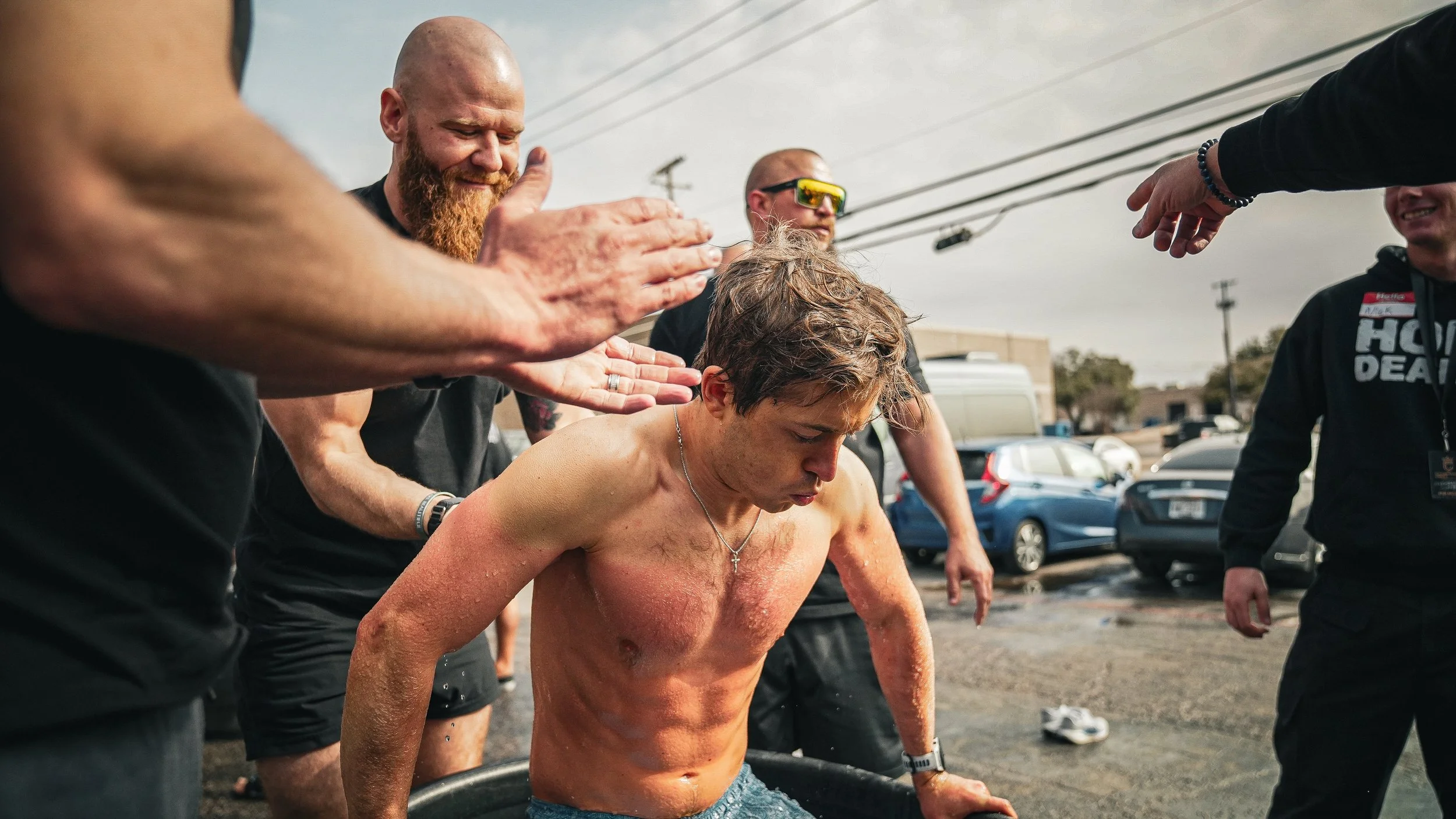 A shirtless young man with wet hair and a crucifix necklace is kneeling in a black baptismal pool as a group of men surround him outdoors on a cloudy day, performing a baptism or spiritual ritual.