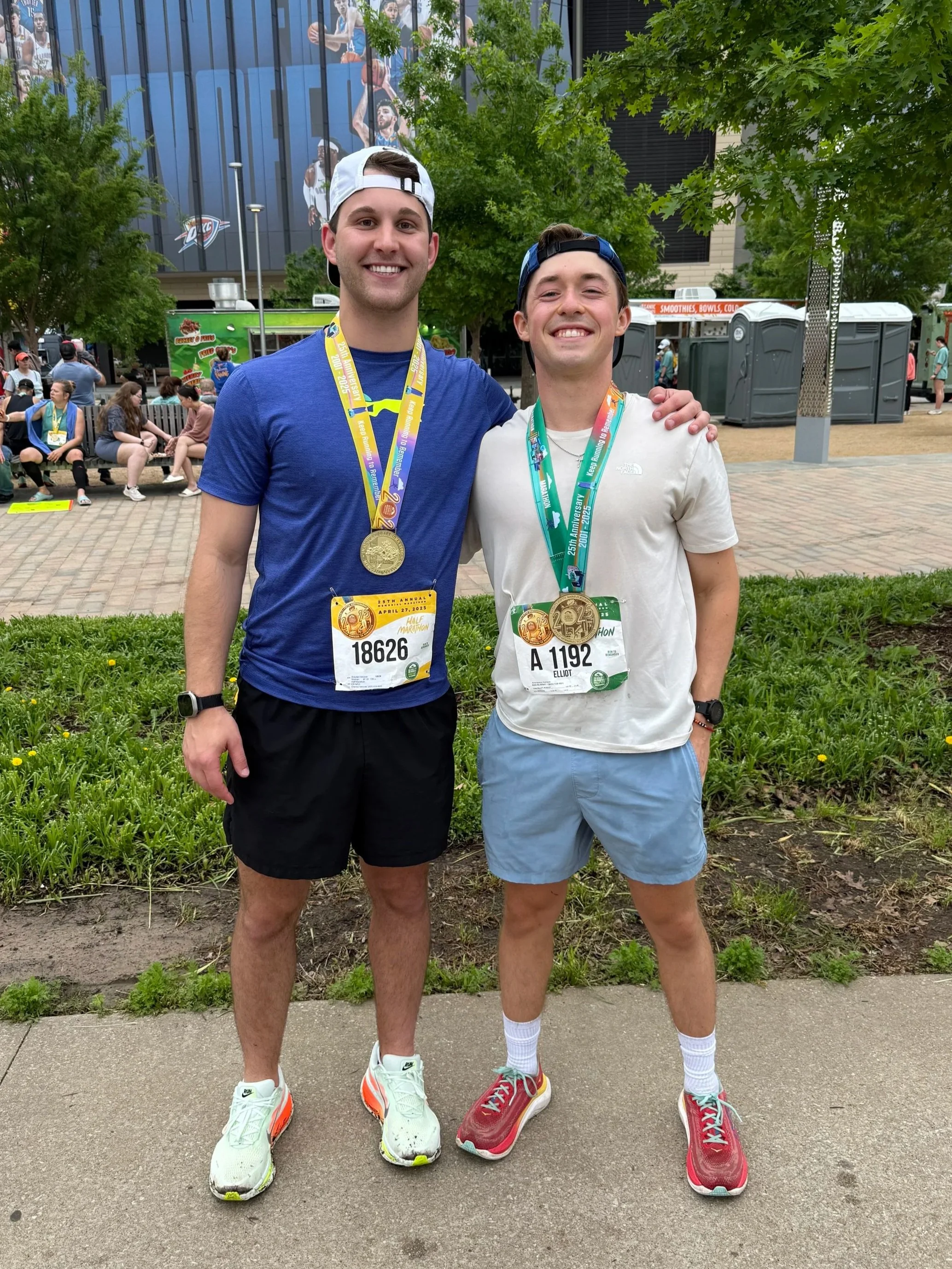 Two young men wearing athletic clothing and medals around their necks pose for a photo outdoors in a park or plaza, smiling. They are standing on a sidewalk near a grassy area with trees, and there are people sitting on benches and walking in the background. Both men have race bibs with numbers, indicating they participated in a race or marathon.