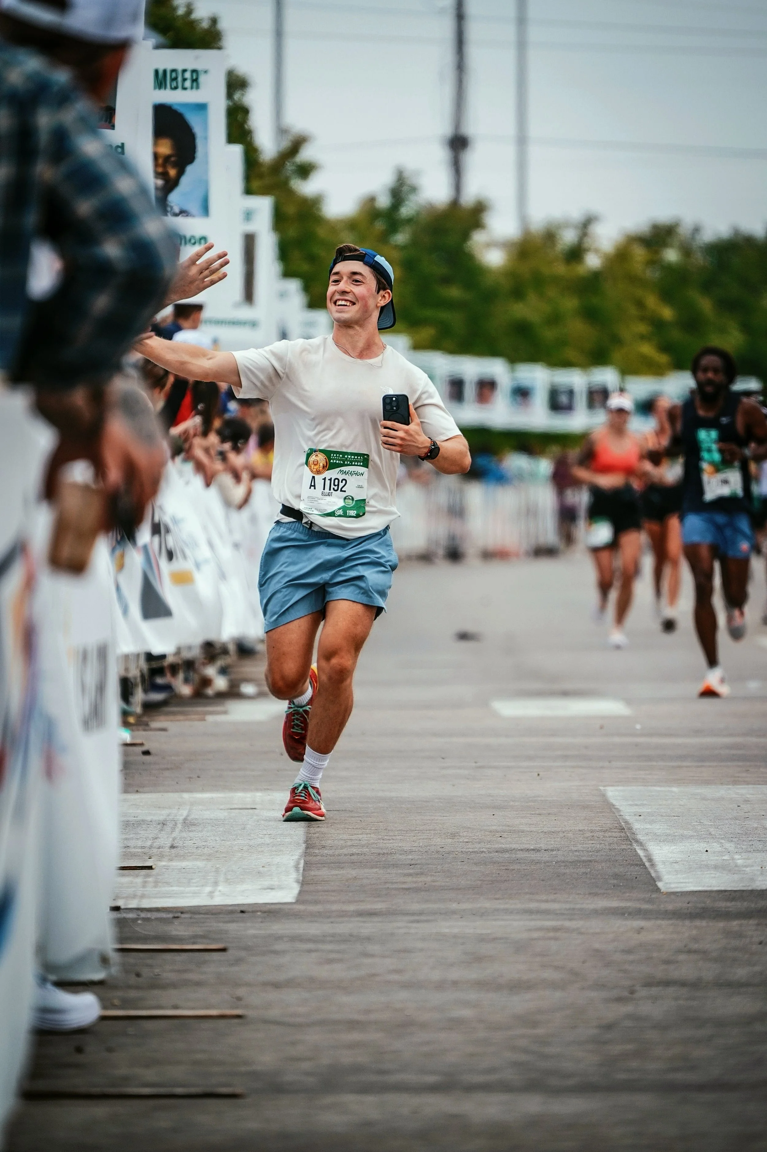 A person running in a marathon, smiling and high-fiving another runner at a race event, with other runners and start banners in the background.