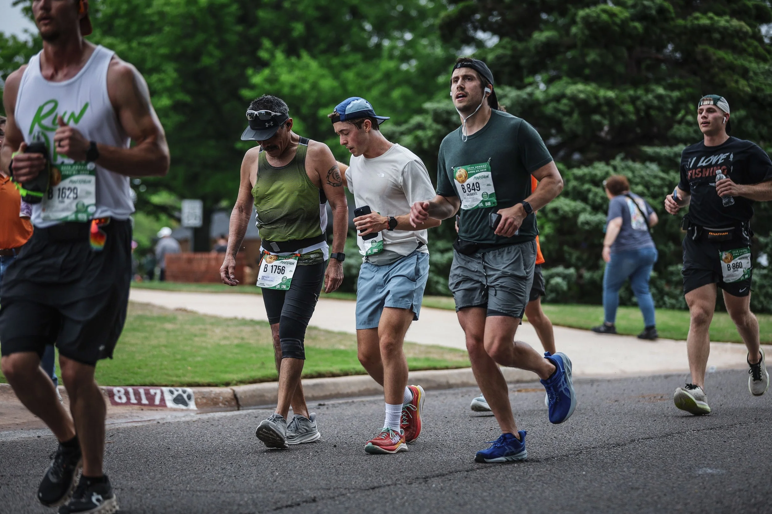 Marathon runners on a paved road in a park, wearing running gear, with bib numbers and some using smartphones.