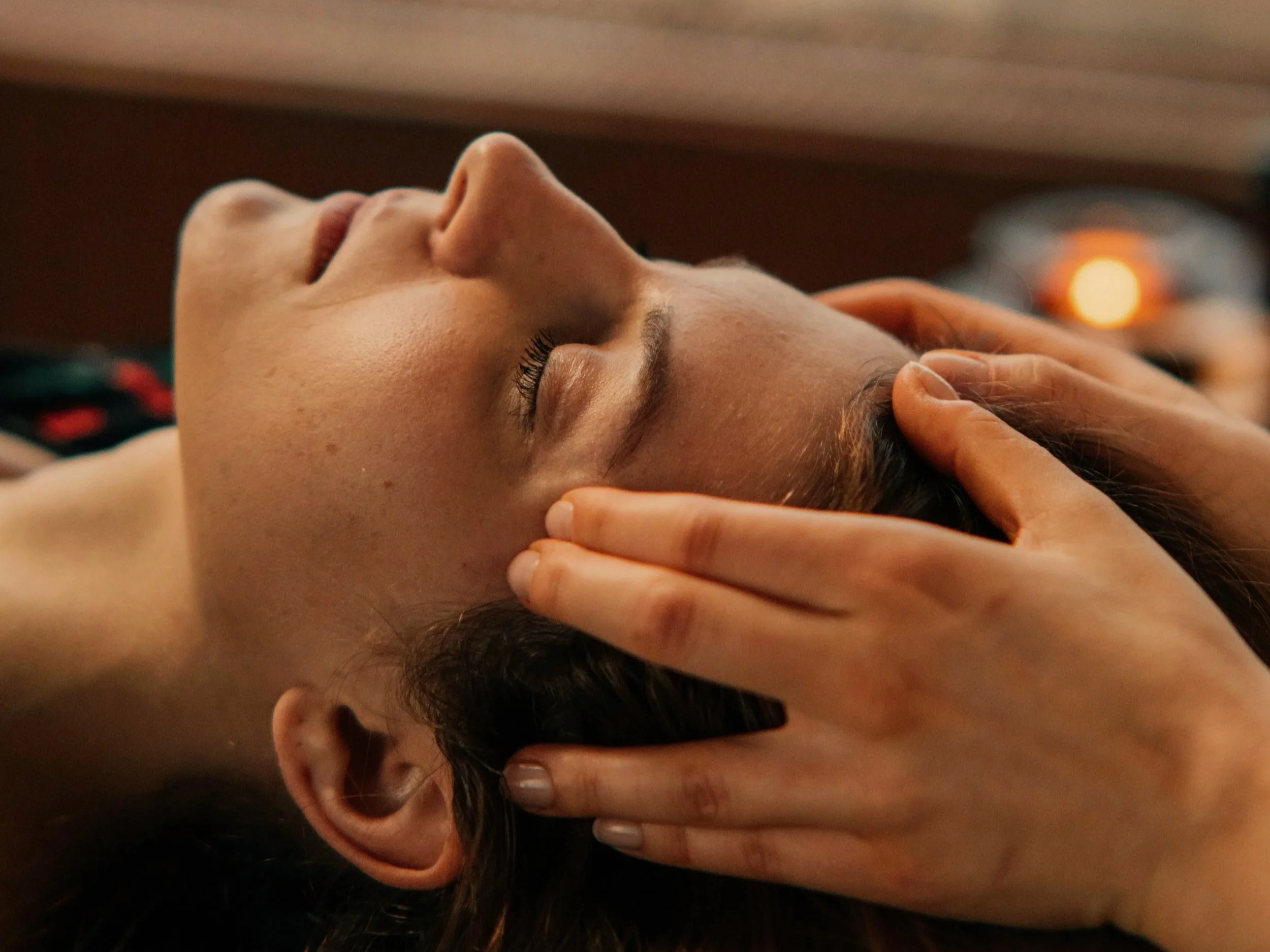 A person receiving a facial massage, lying down with eyes closed, while a therapist gently presses temples with fingers.