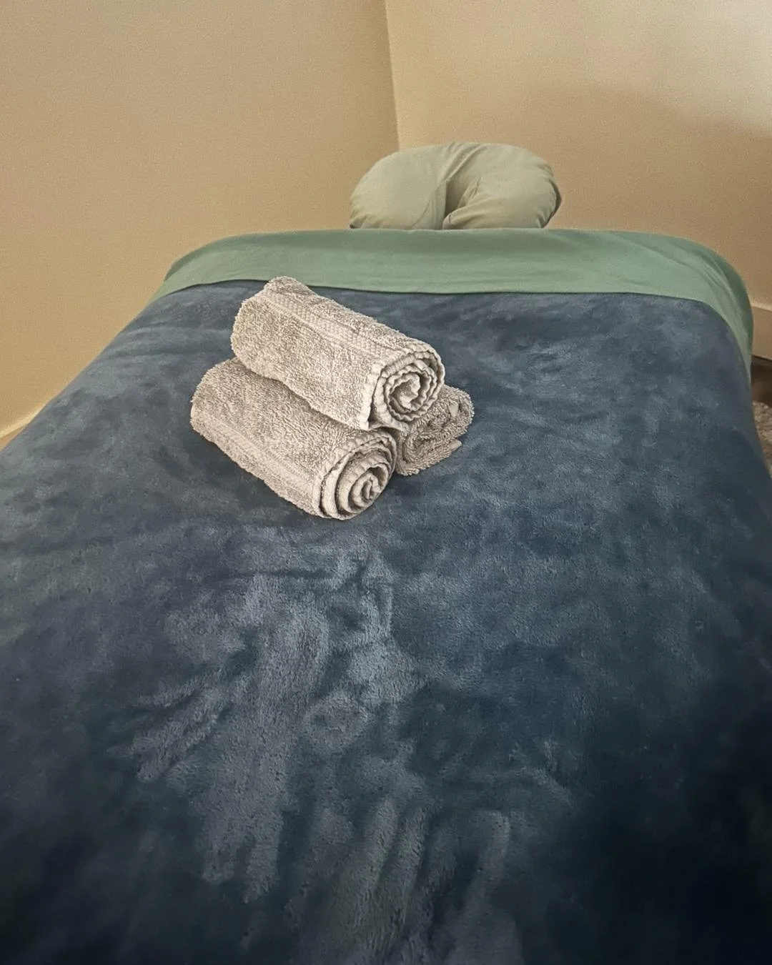 A neatly made massage table with a blue plush blanket, a green sheet, a green pillow at the head of the bed, and three rolled gray towels placed on the blanket in a soothing massage room