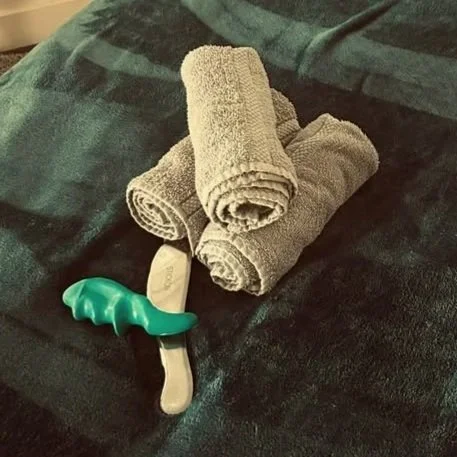 Three rolled gray towels placed on a dark surface next to a green massage tool and a silver gua sha tool