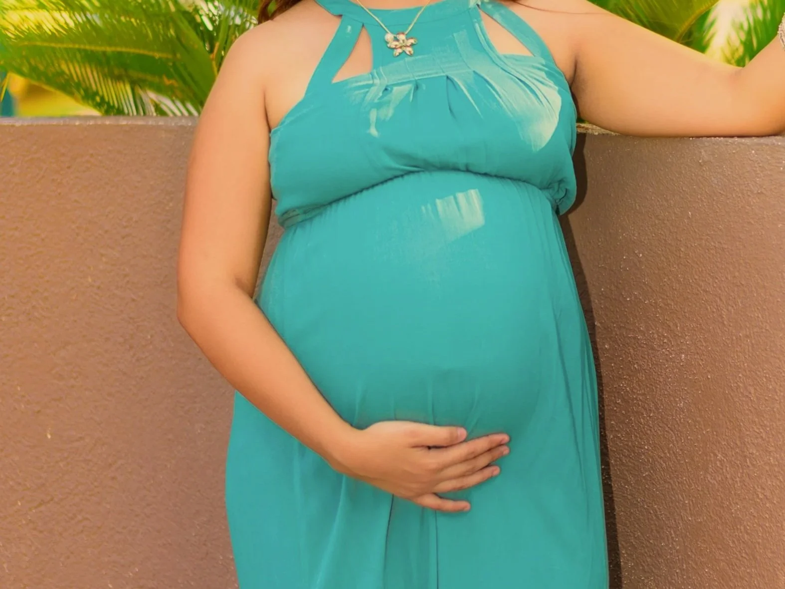 A pregnant woman wearing a teal sleeveless dress and a butterfly pendant necklace, standing against a brown wall with green tropical leaves in the background, gently holding her baby bump with one hand.