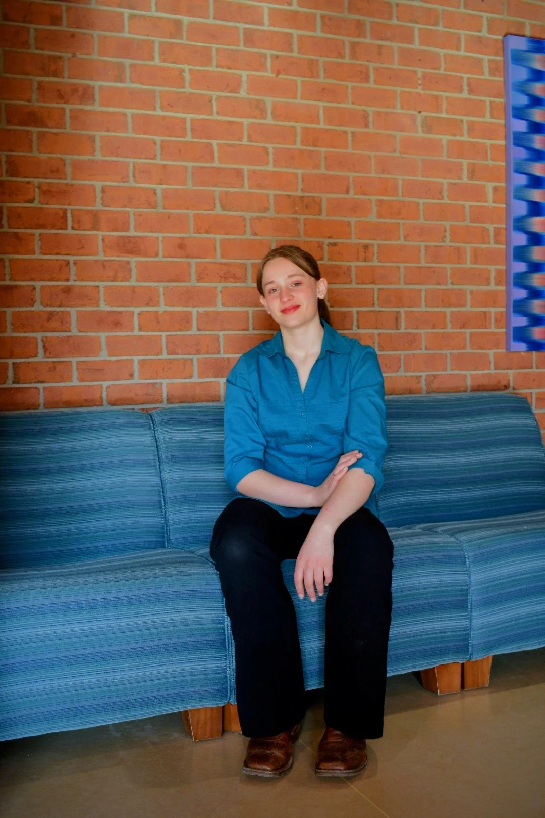 A young woman with red hair, wearing a blue button-up shirt and black pants, sitting on a blue striped sofa with her arms crossed, in front of a brick wall.