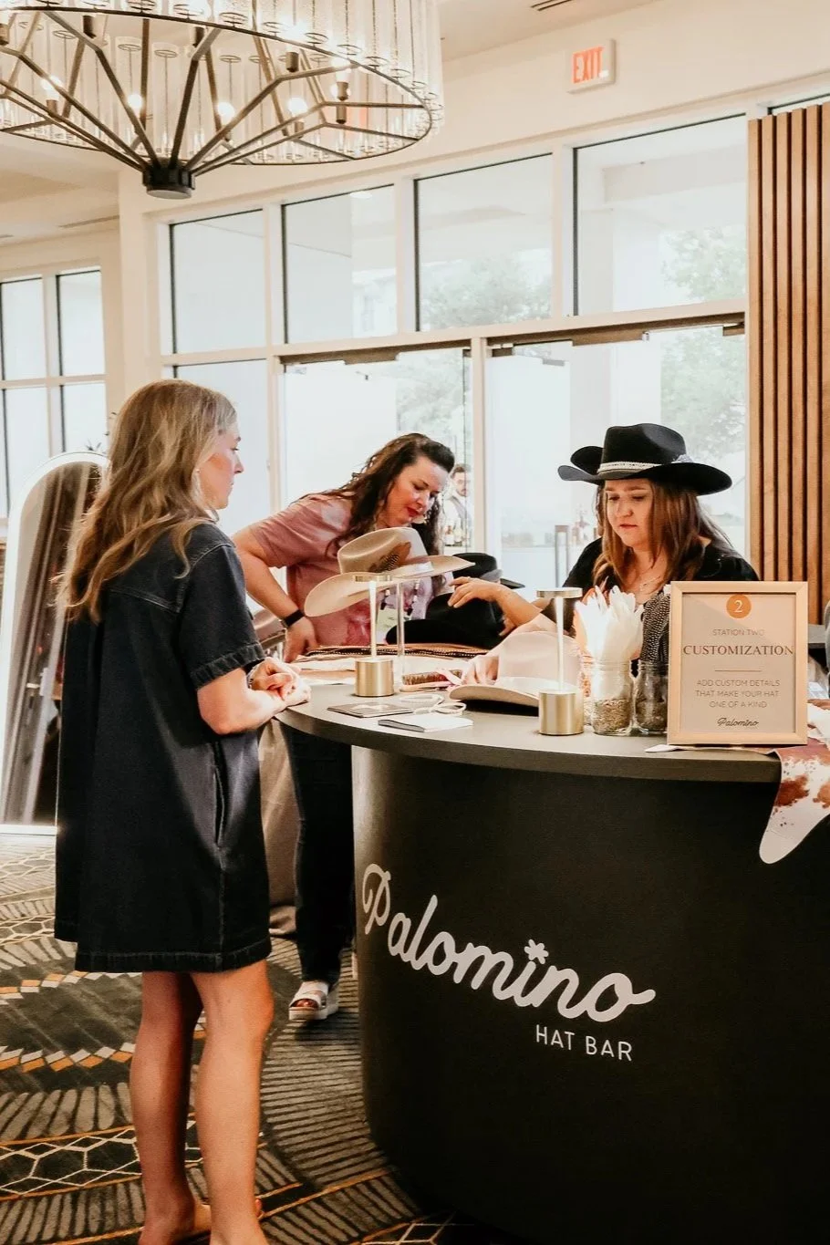 Three women at a hat customization station at Palomino Hat Bar, with large hats and a sign indicating customization options.