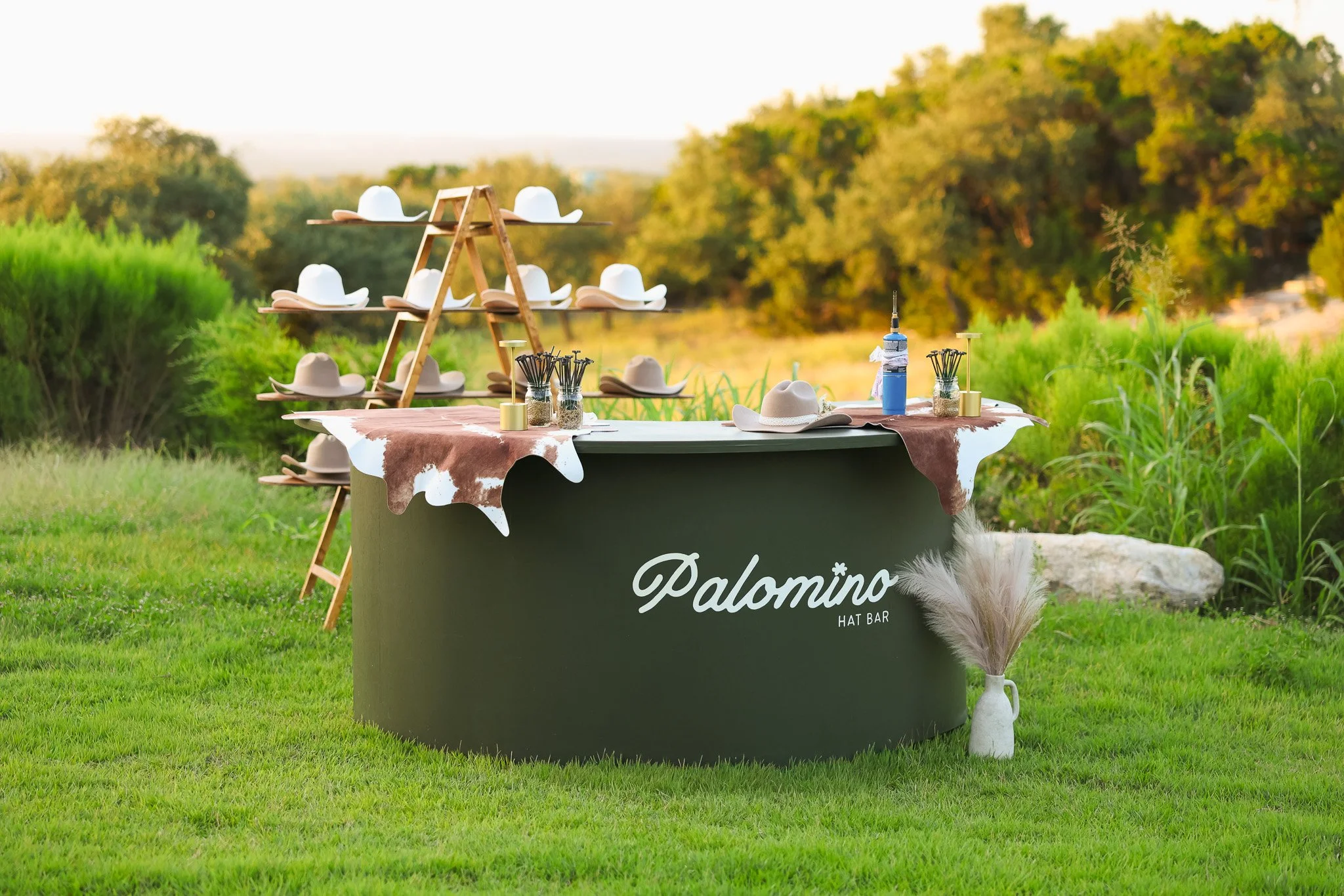 Outdoor hat bar display with white cowboy hats on wooden shelves, set on a table with jars of decorations, in a grassy area with green trees in the background.