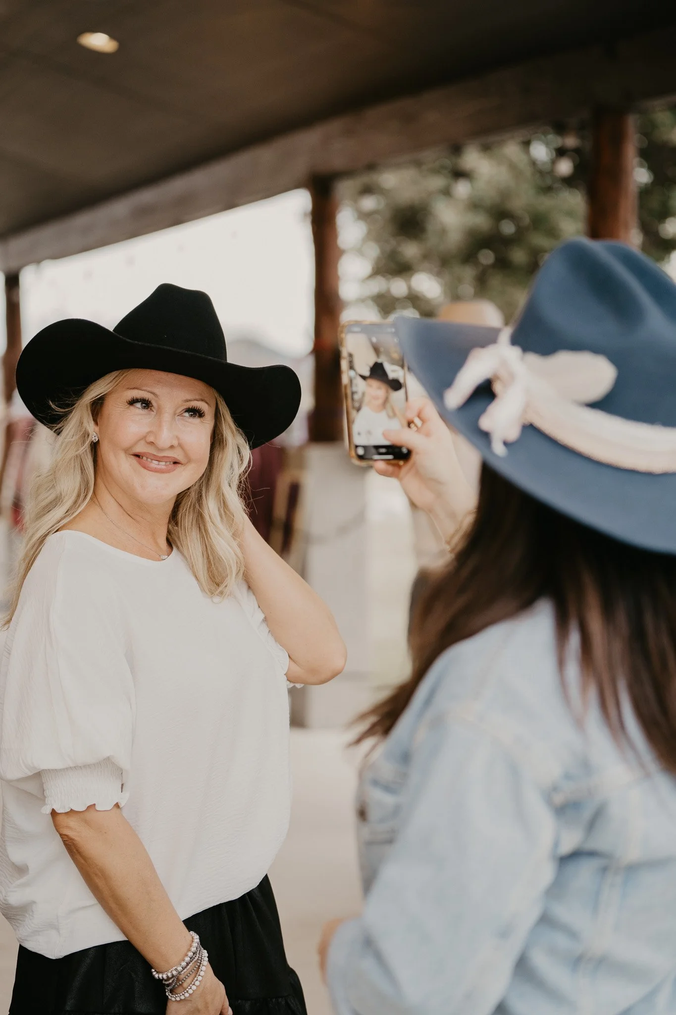 Woman with blonde hair wearing a black hat and white blouse smiling for a photo taken by someone wearing a blue hat with a decorative ribbon. The background is blurred with indoor and outdoor elements.
