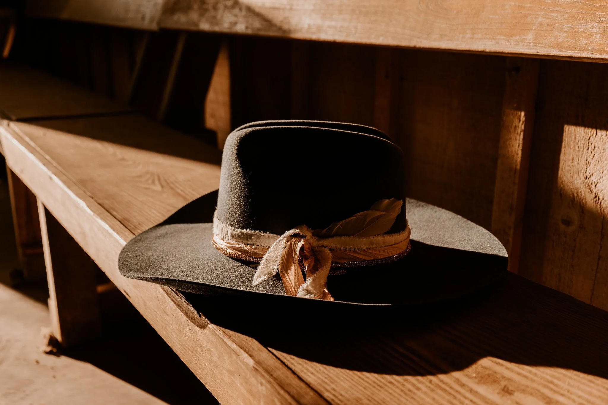 A black felt hat with a beige ribbon and feather, resting on a dark surface in front of a wooden backdrop.