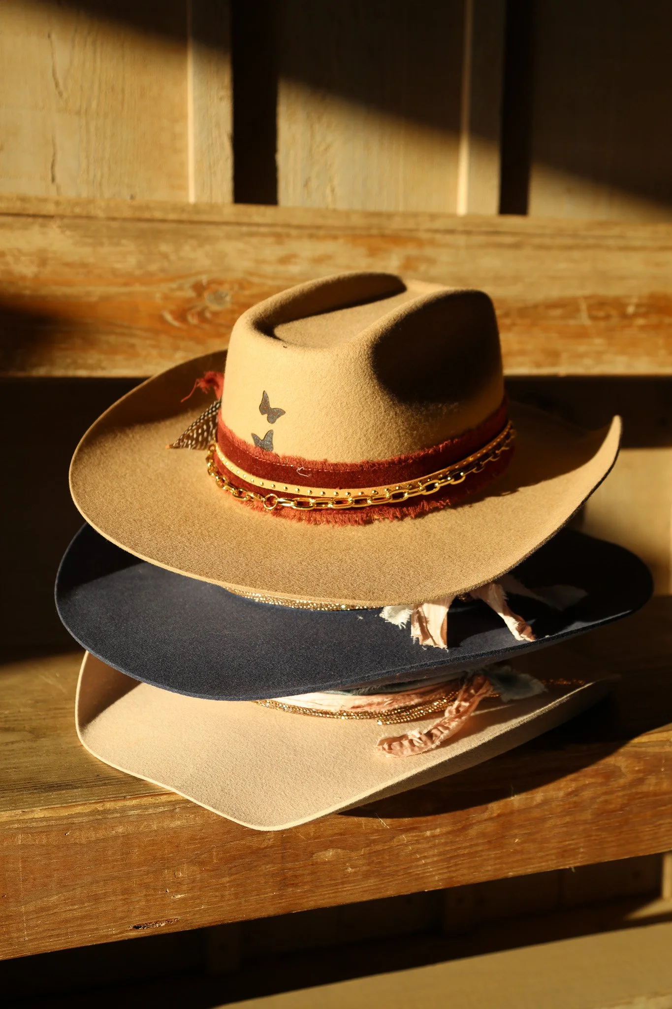 Stacked cowboy hats on a wooden surface, featuring various colors and decorative elements like chains and ribbons, with natural sunlight casting shadows.