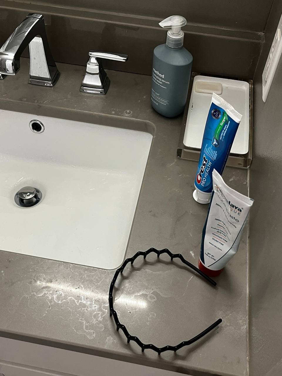 Bathroom sink countertop with a black headband, a gray soap dispenser, a small tray, and two tubes of toothpaste.