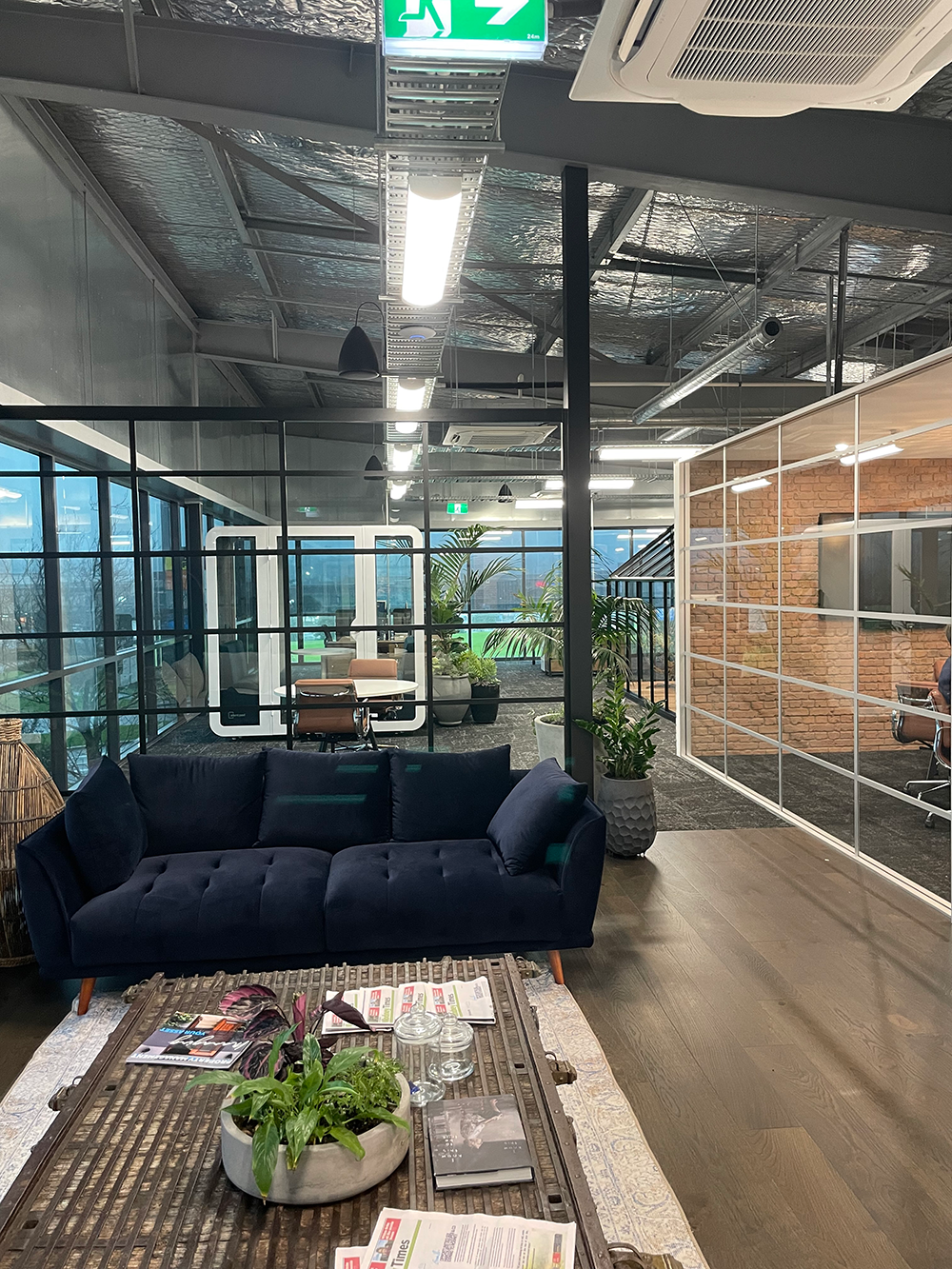 Modern office lounge with a black sofa, indoor plants, a glass coffee table with magazines and decorative items, and subdivided workspaces with glass and brick walls.
