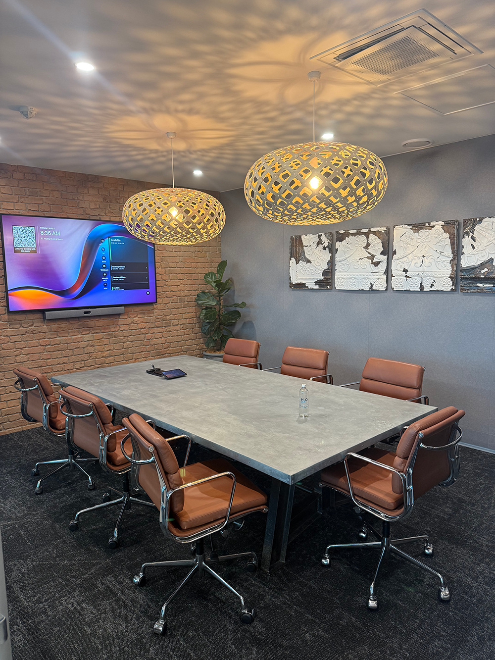 A conference room with a large rectangular table, eight leather chairs, a wall-mounted screen, decorative artwork on the wall, hanging pendant lights, and a potted plant in the corner.