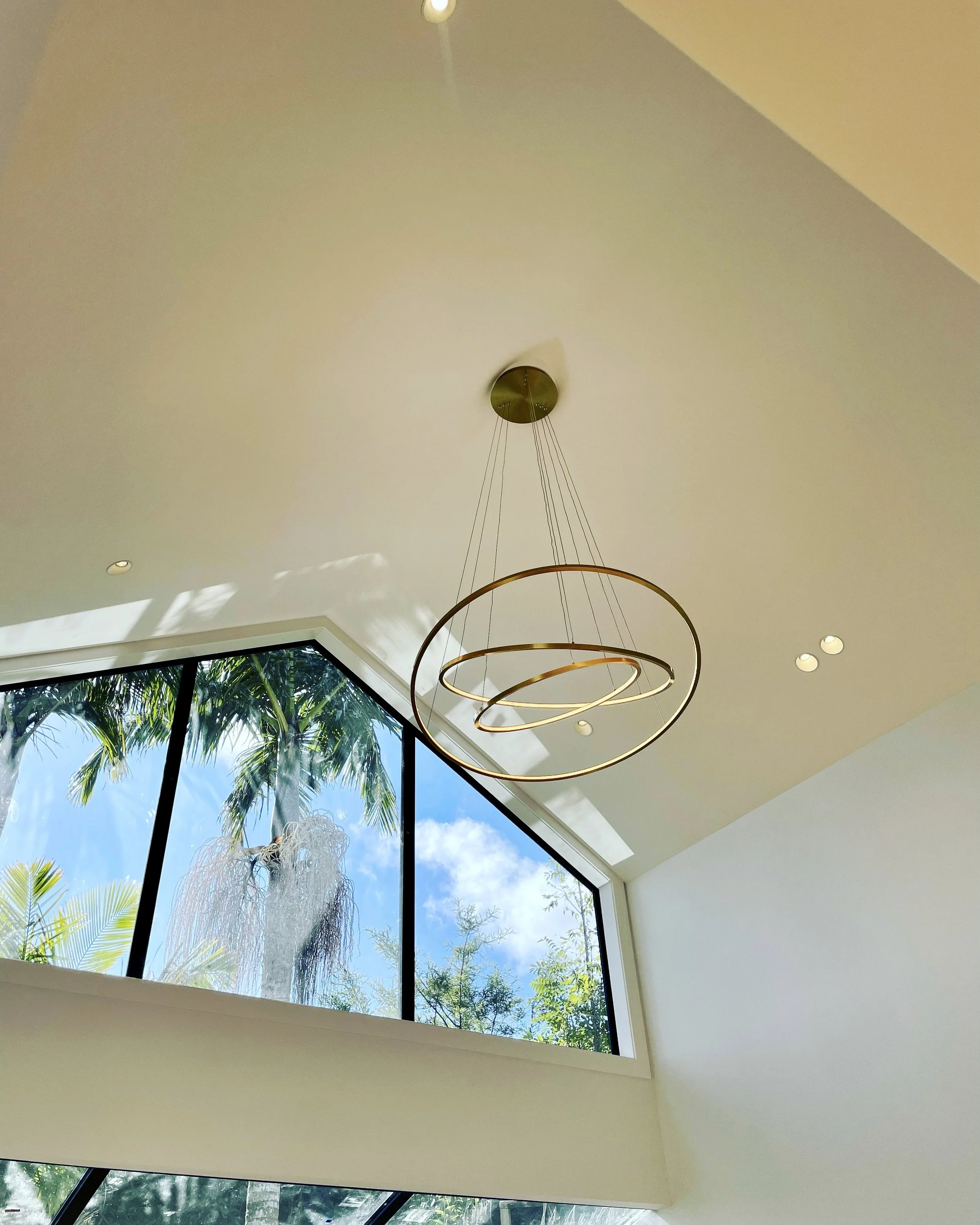 Modern interior view with a large window showing palm trees outside, a circular chandelier hanging from the ceiling, and recessed ceiling lights.