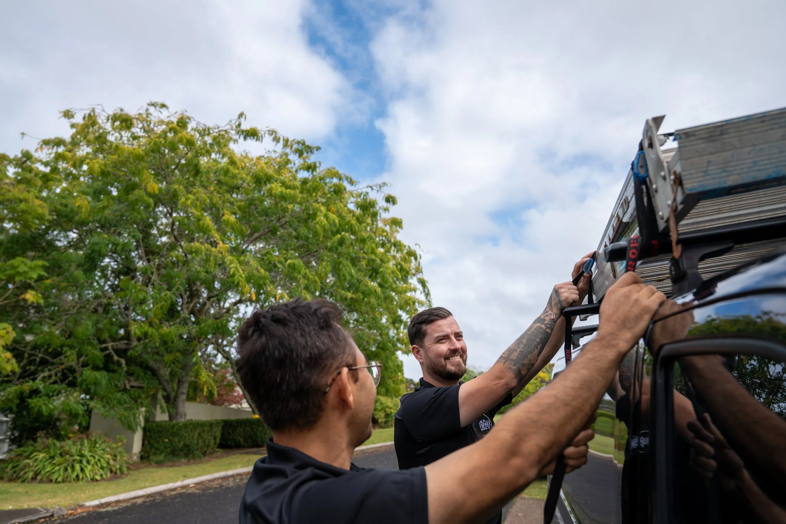 Group of people working together outdoors, installing or adjusting equipment on a vehicle roof, with trees and cloudy sky in the background.