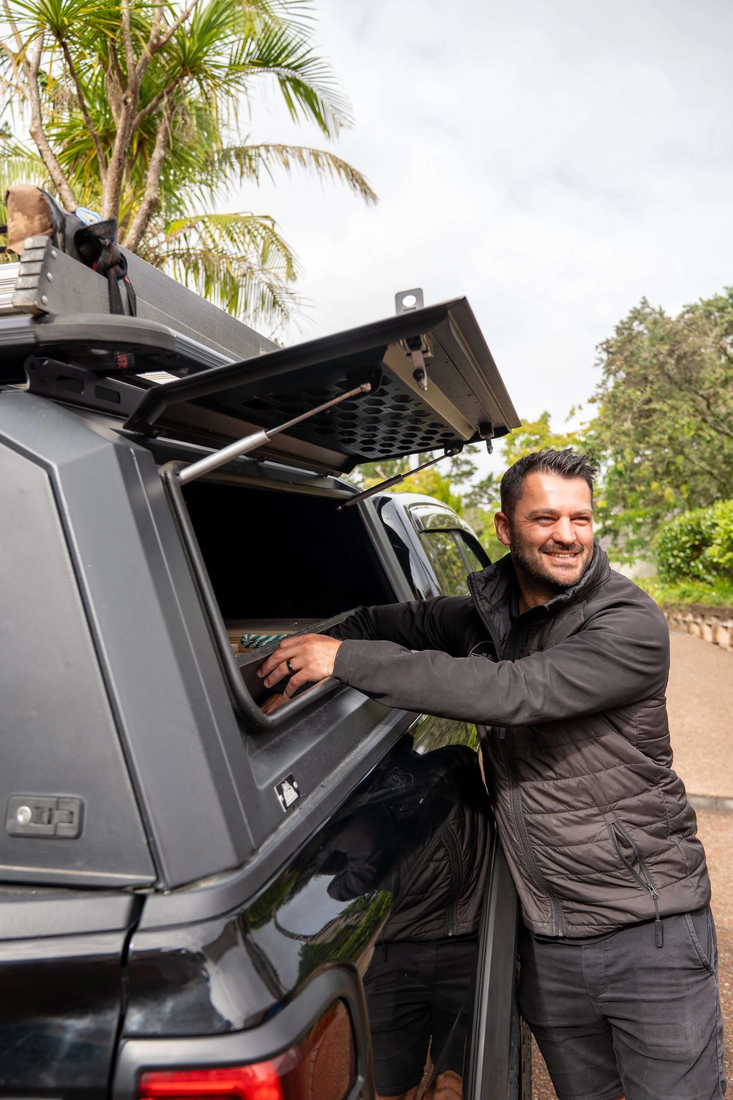 A man with short dark hair and a beard is smiling while leaning out of the open window of a black SUV. He is wearing a black jacket. The SUV has a rooftop storage box, and there are palm trees and other greenery in the background under a cloudy sky.