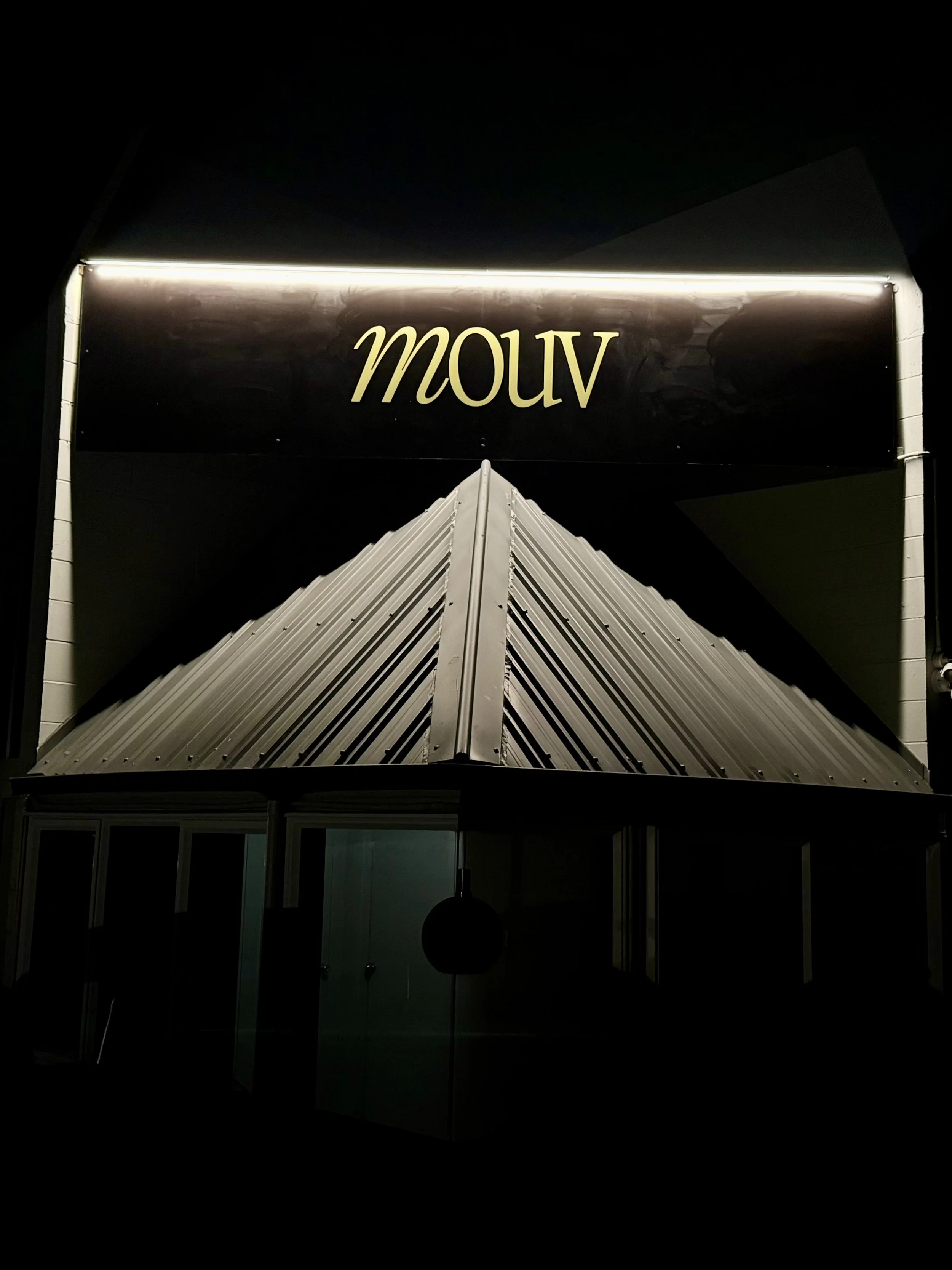 Nighttime view of a sign that reads 'mouv' in illuminated letters above a building entrance with a sloped metal roof.