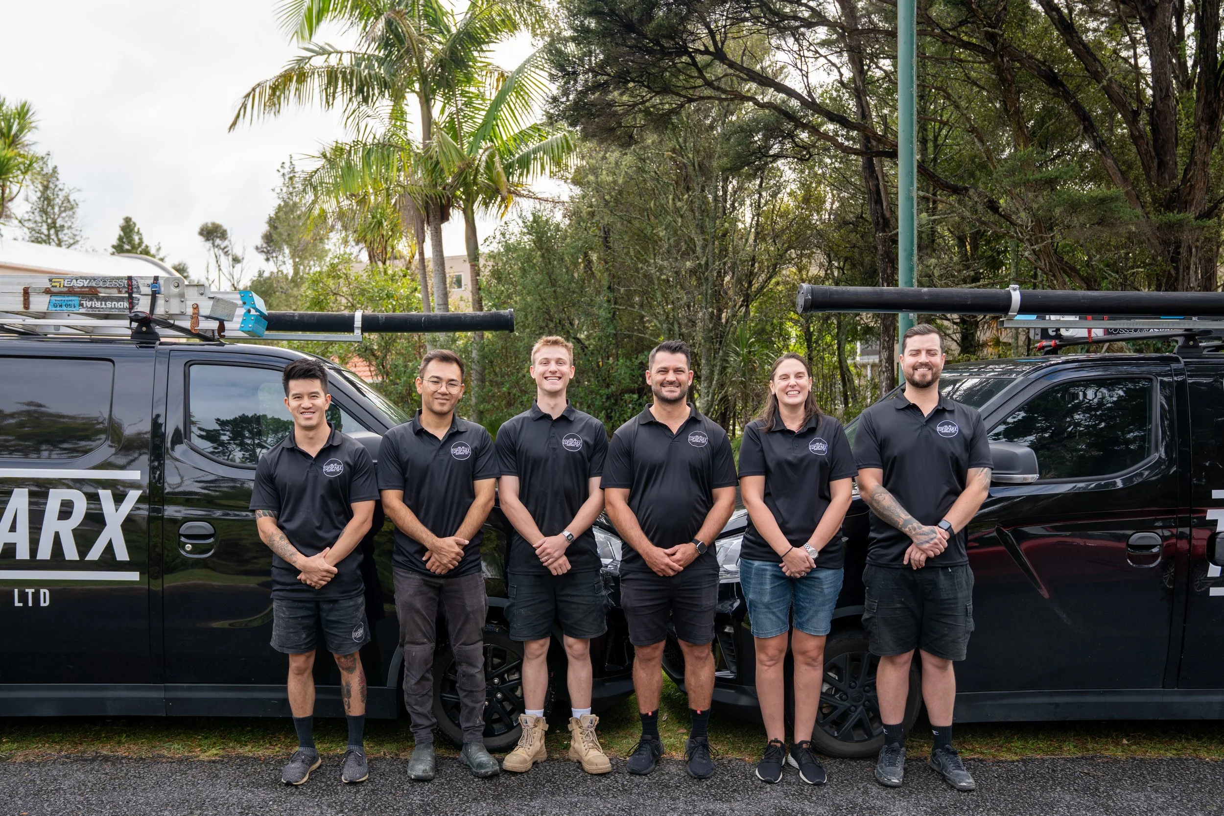 A group of six employees standing outdoors in front of black company vehicles, smiling. They are wearing black uniforms with company logos, surrounded by trees and palm trees.