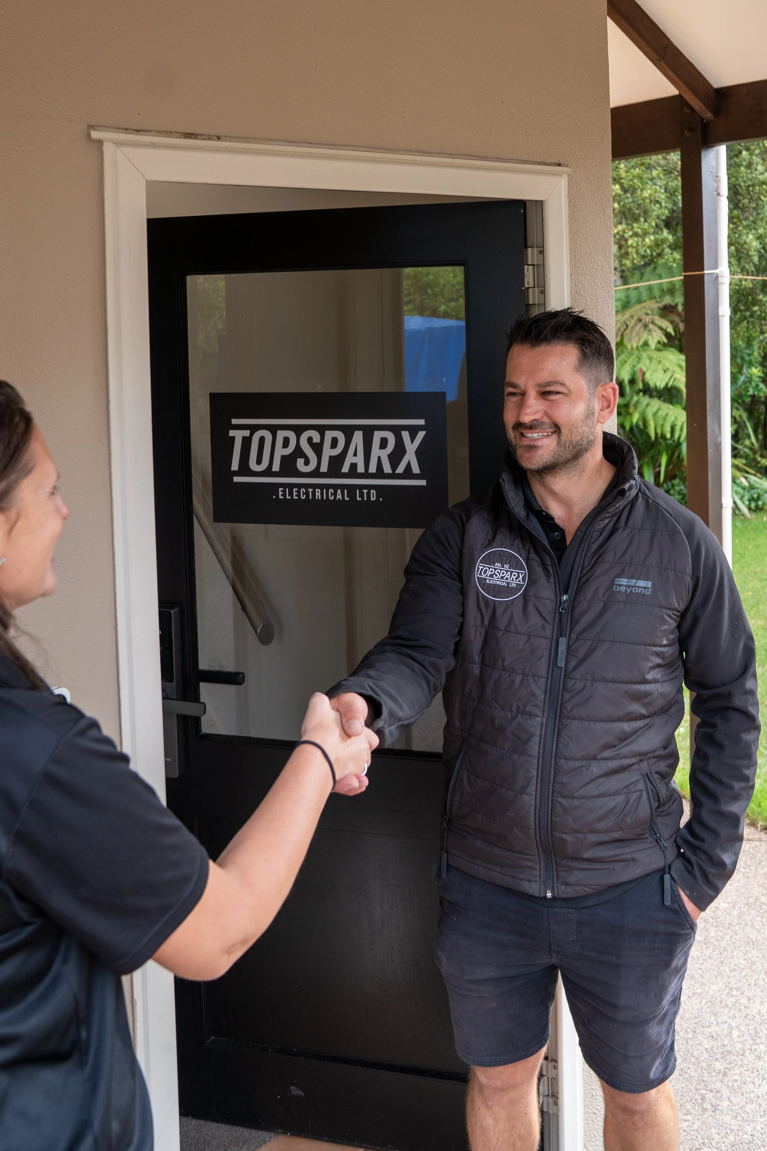 Two people shaking hands outside in front of a door with a sign that reads "TOPSPARX ELECTRICAL LTD." One person is a man wearing a dark jacket and shorts, smiling. The other person's face is not visible.