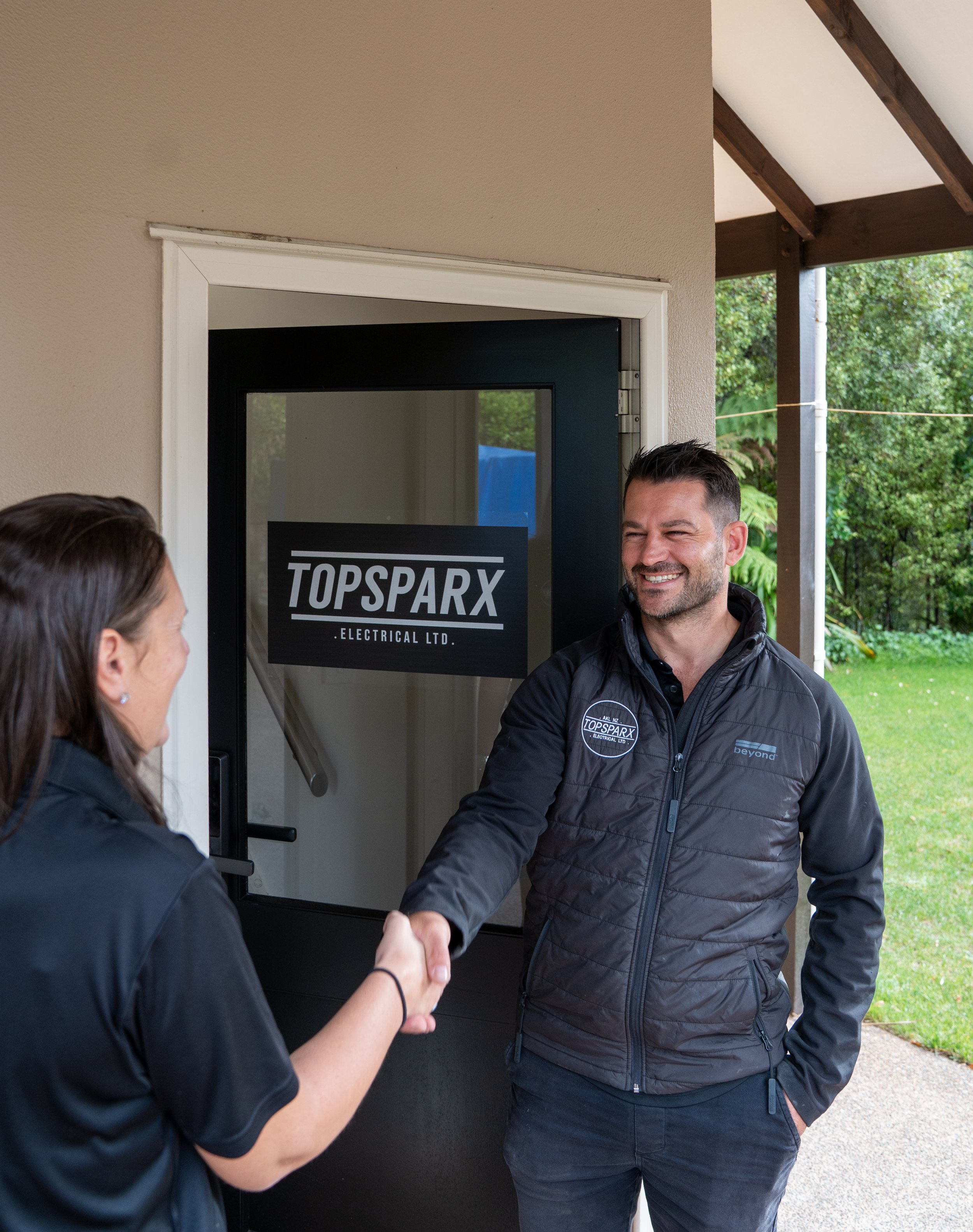 Two people shaking hands outside a building with a sign that reads 'TOPSPARX ELECTRICAL LTD.' The man is smiling and wearing a black jacket with the company logo. The woman has dark hair and is wearing a dark jacket.