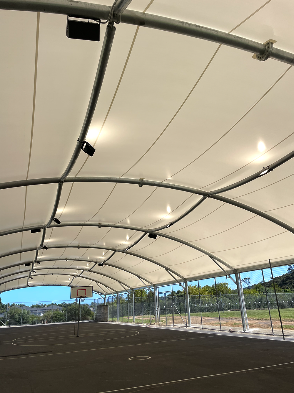 Indoor basketball court with a high, curved ceiling, basketball hoop, and a netted outer wall with trees outside.