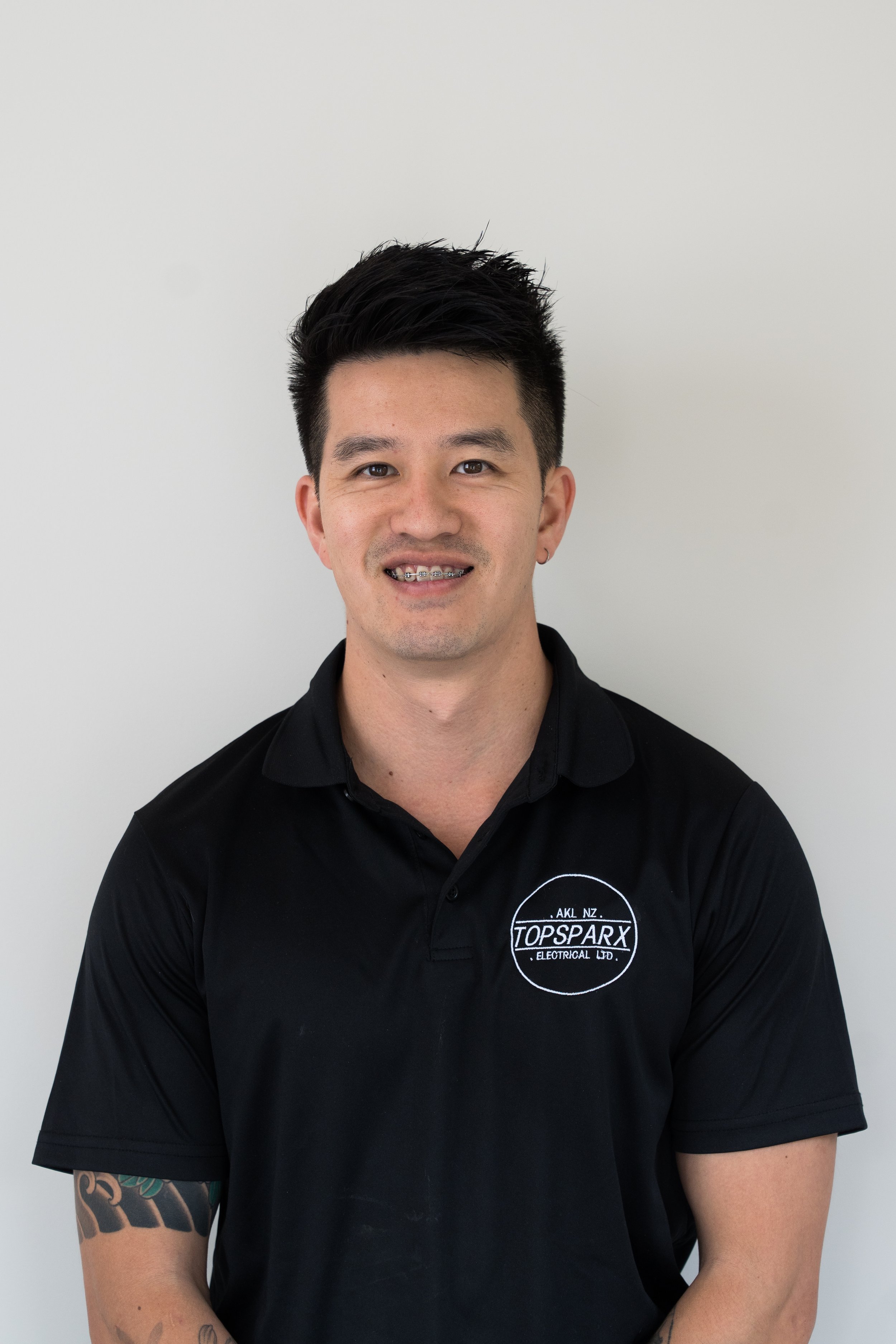 Portrait of a young man with black hair styled up, wearing earrings and braces, in a black shirt with a logo for TOPSPARX Electrical Ltd., standing against a plain white background.