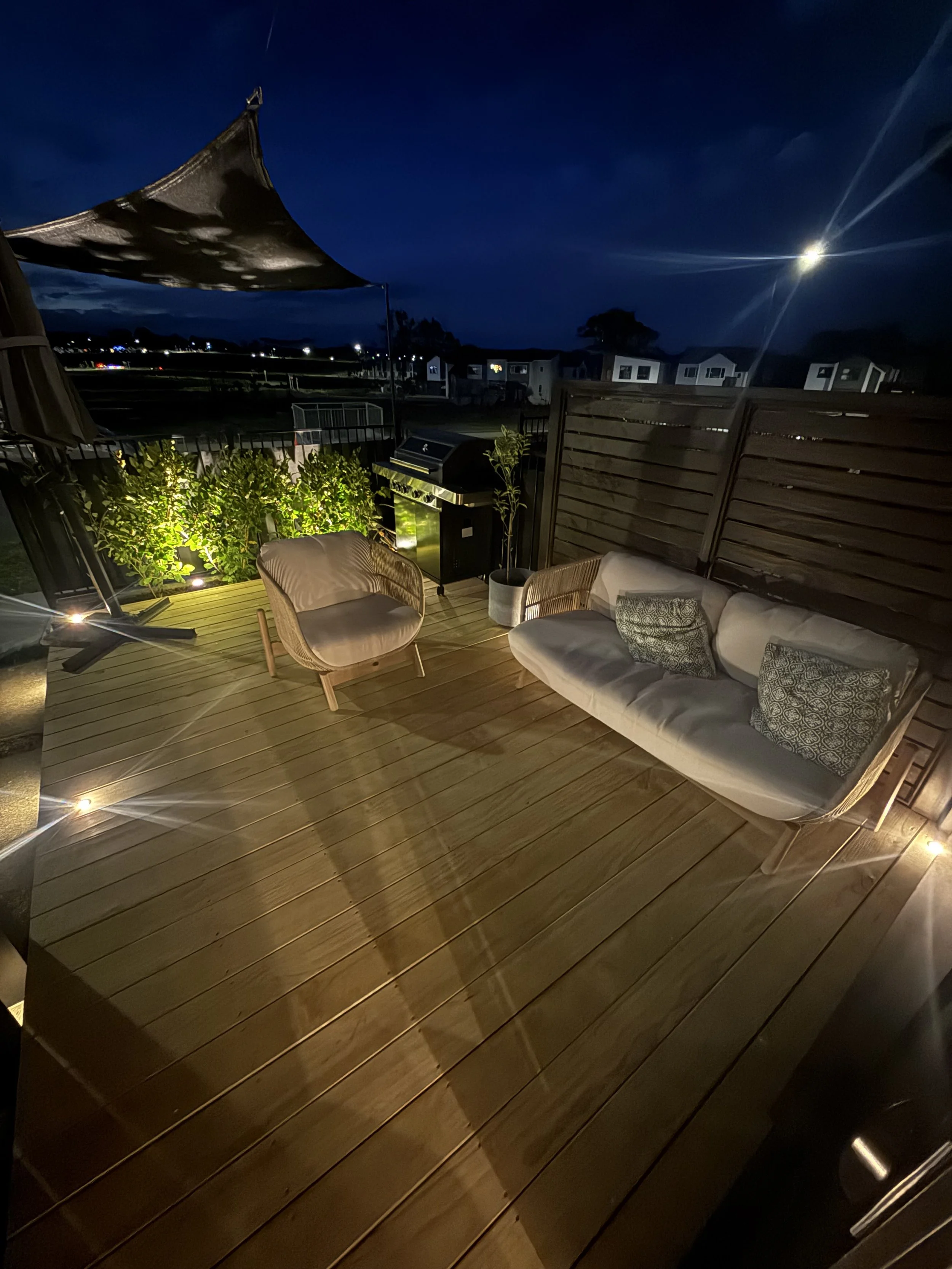 Nighttime view of a backyard patio with a wooden deck, a beige cushioned outdoor sofa with patterned pillows, a wicker chair, a barbecue grill, and greenery, with a dark sky, bright moon, and nearby houses.