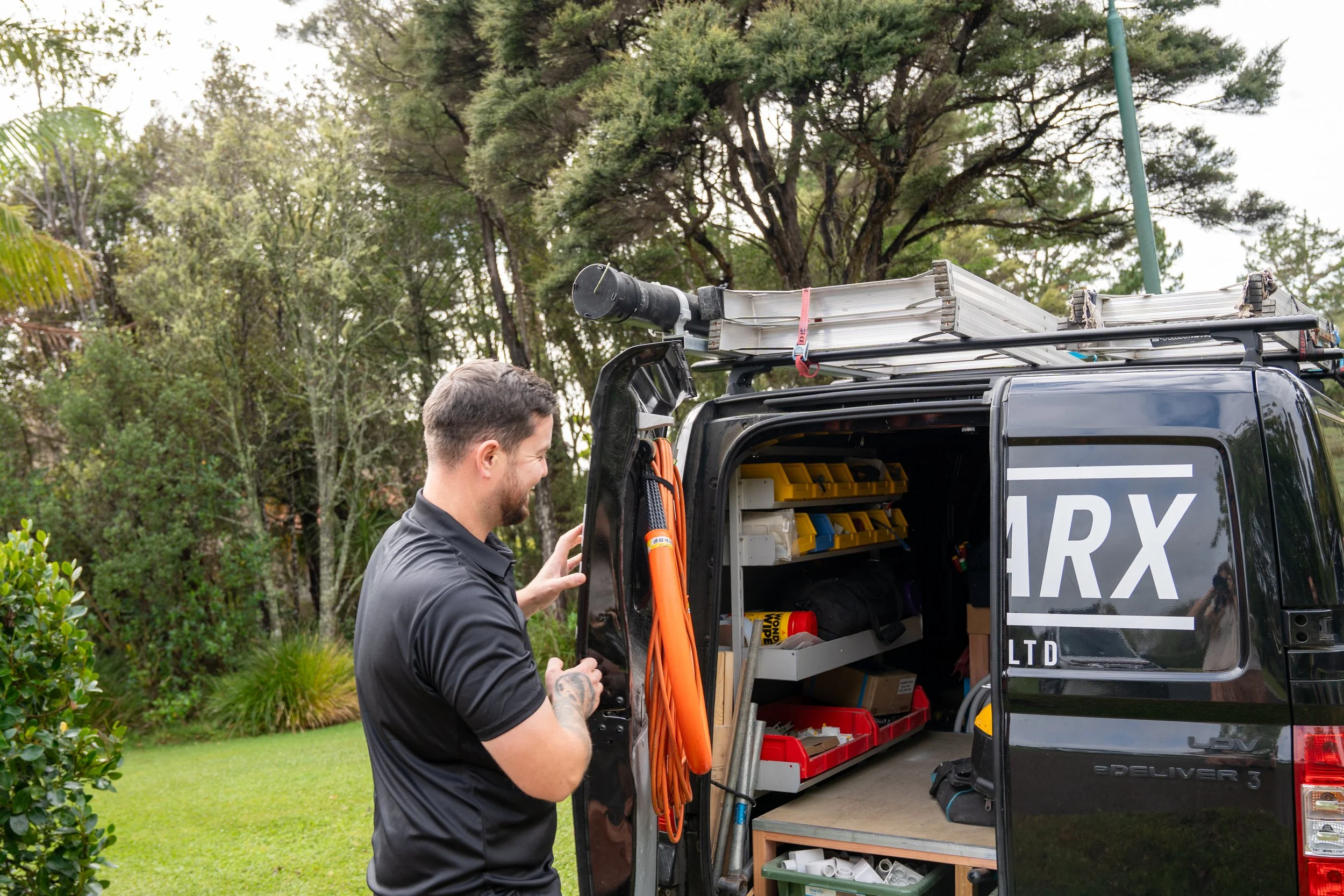 A man with tattoos working near an open utility vehicle filled with tools and equipment outdoors, with trees and greenery in the background.