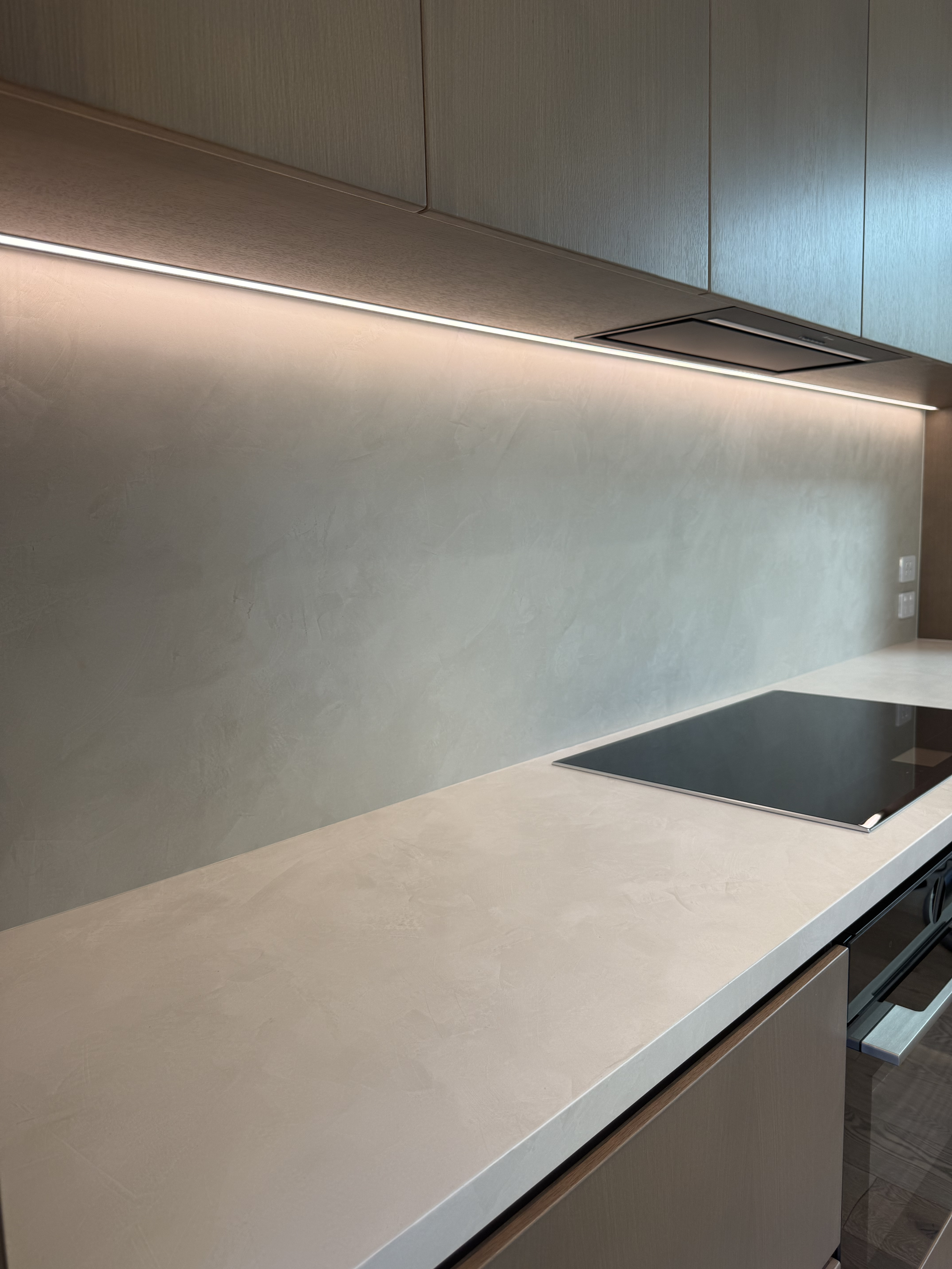 Modern kitchen countertop with a sleek, black induction cooktop and beige textured backsplash, illuminated by under-cabinet lighting.