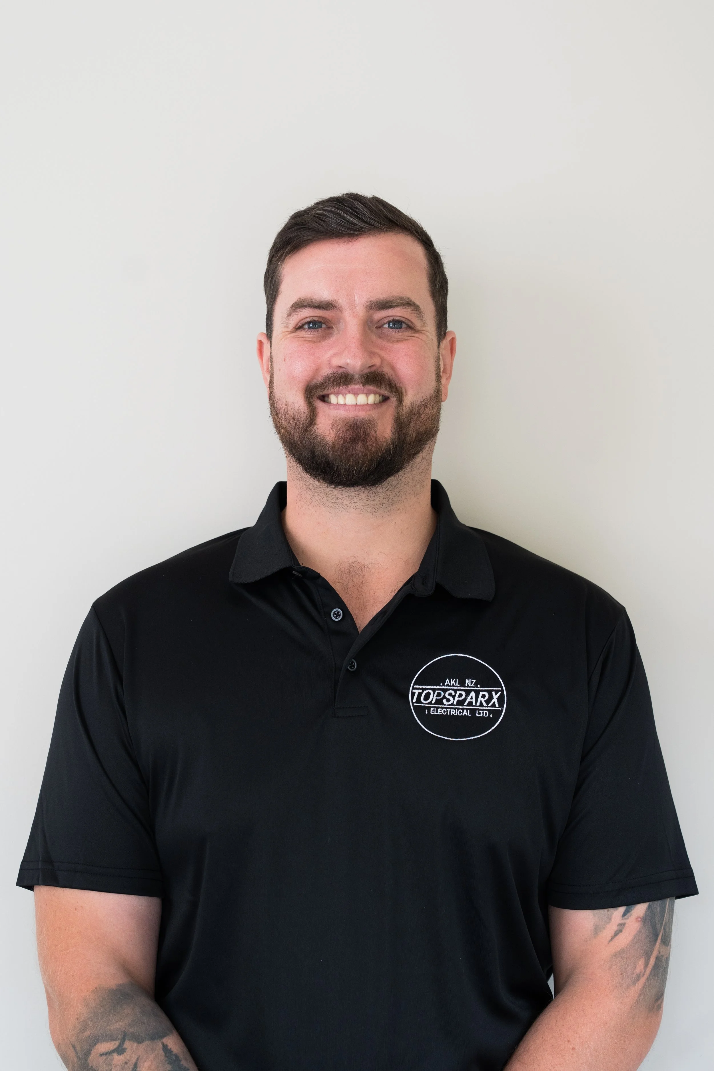 Two men are loading a wooden ladder onto a black van parked on a street in a suburban neighborhood with trees and cars in the background. One man is smiling and holding the ladder while the other is taking a photo.