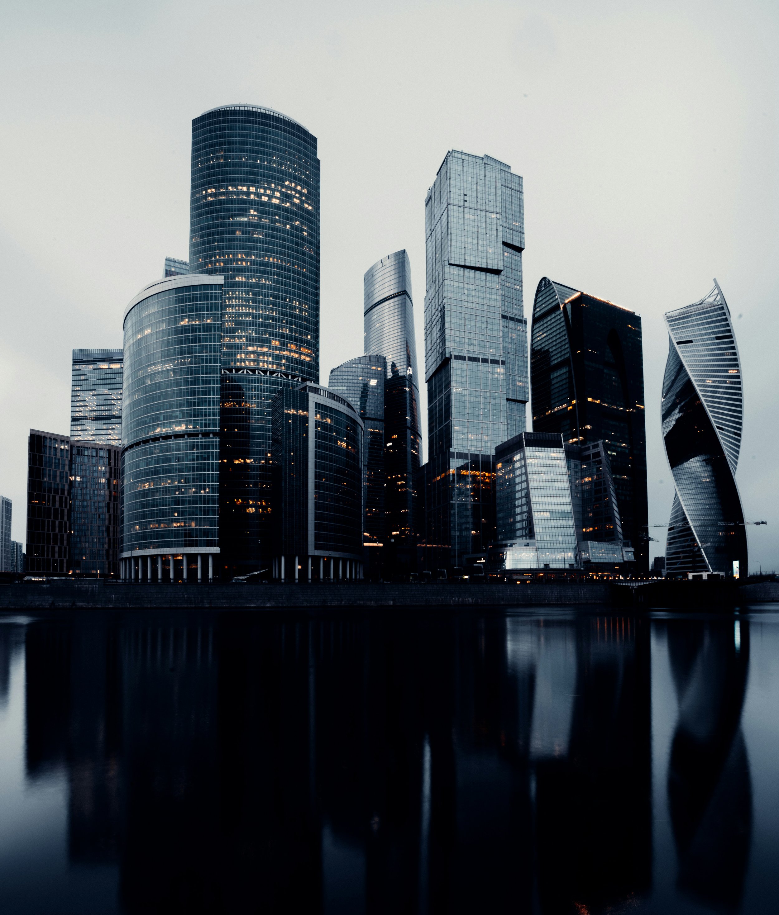 Nighttime cityscape with tall modern skyscrapers reflecting in a river.
