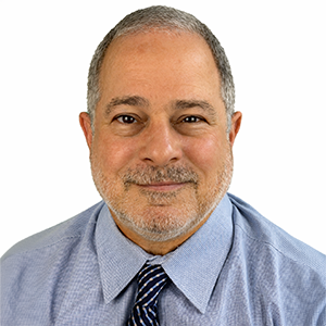 Close-up portrait of a middle-aged man with short gray hair and a beard, wearing a light blue shirt and a striped tie, smiling against a plain white background.