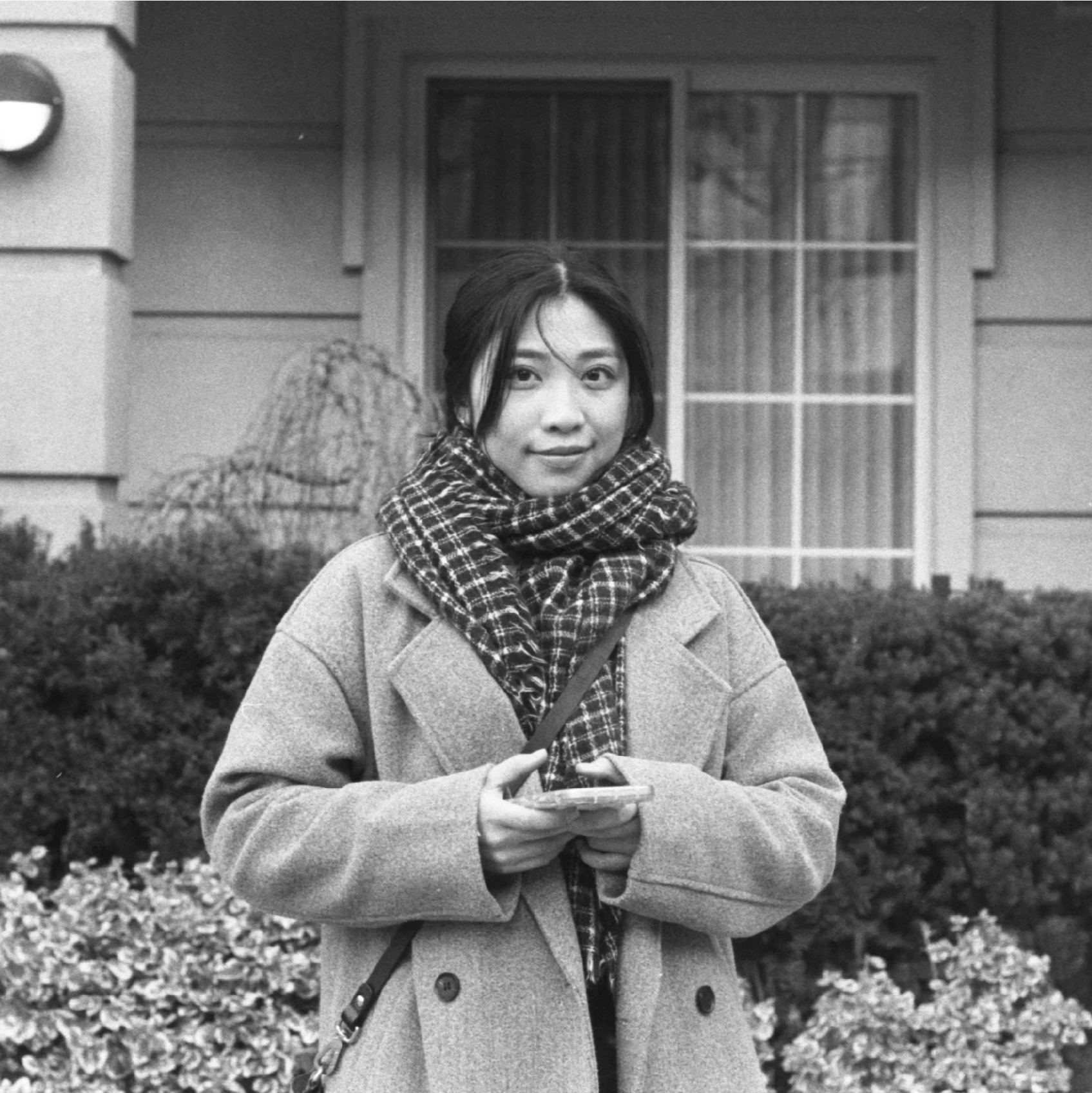 A young woman with dark hair, wearing a patterned scarf and a coat, standing outdoors in front of a house with large windows, holding a smartphone.