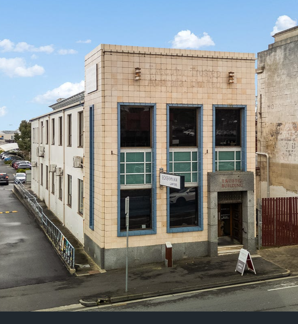 A small, three-story building with beige tiles on the front and large dark windows. Signs read "Eckermann Lawyers" and "Trustee Building." The building is on a city street with parked cars and a sidewalk.