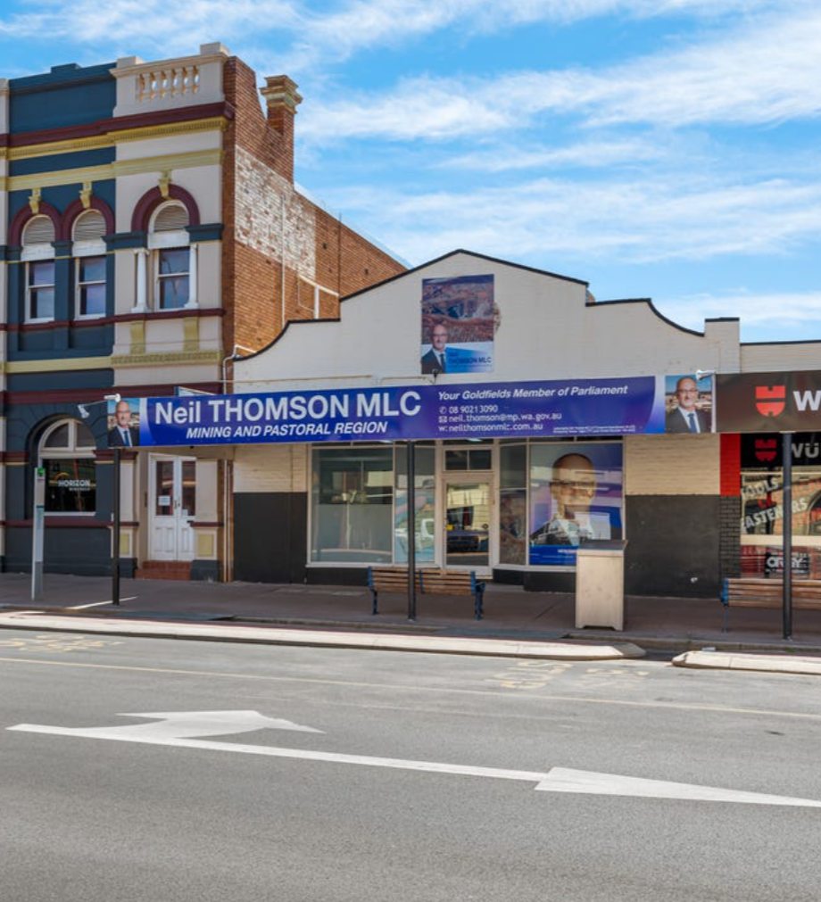 A building with campaign signs for Neil Thomson and political posters, with a blue sky and some clouds in the background. There are street benches and a bus stop in front of the building.