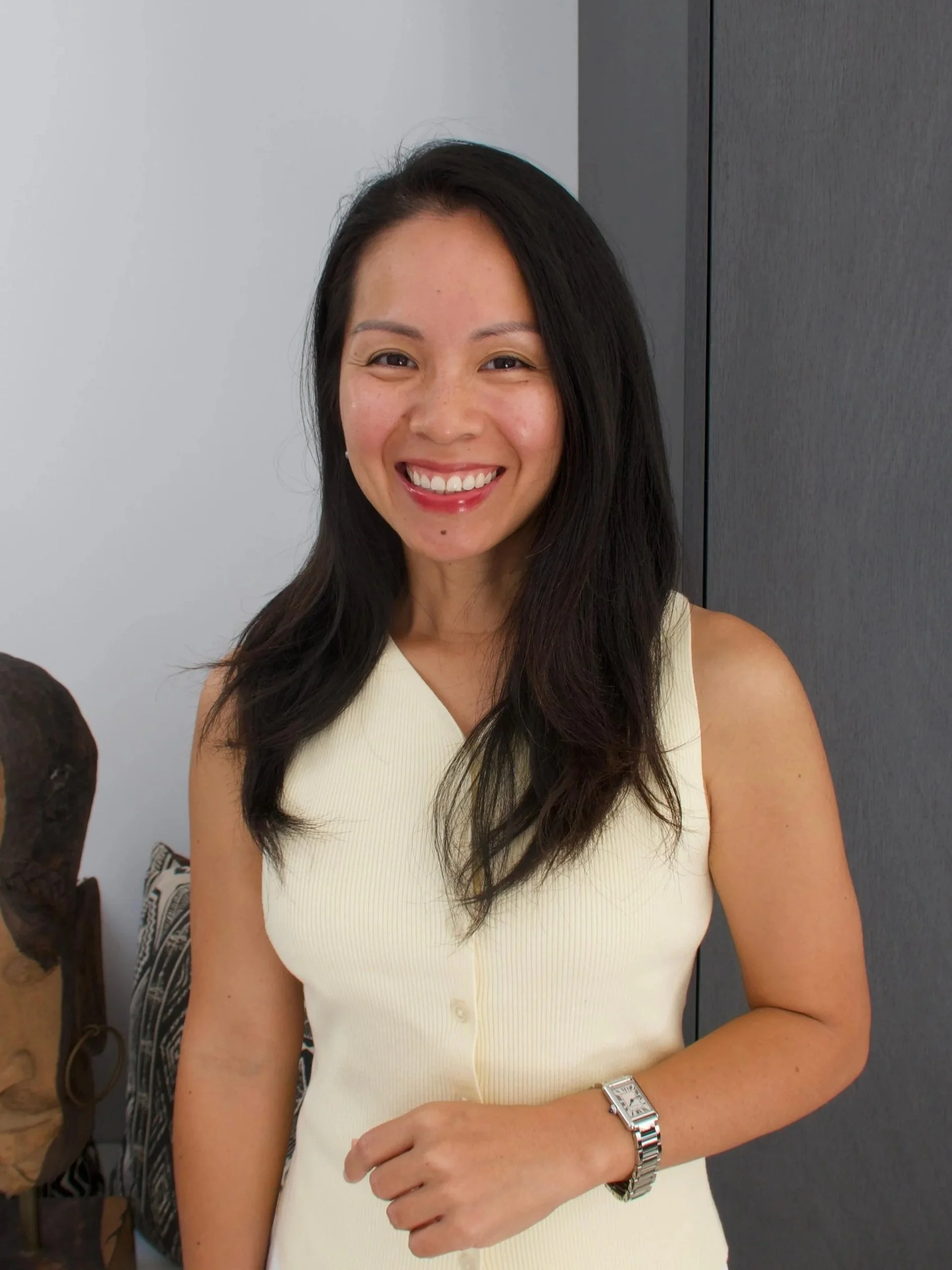 A woman with long dark hair, smiling, wearing a sleeveless cream-colored top, and a silver watch on her left wrist, standing in front of a neutral background.