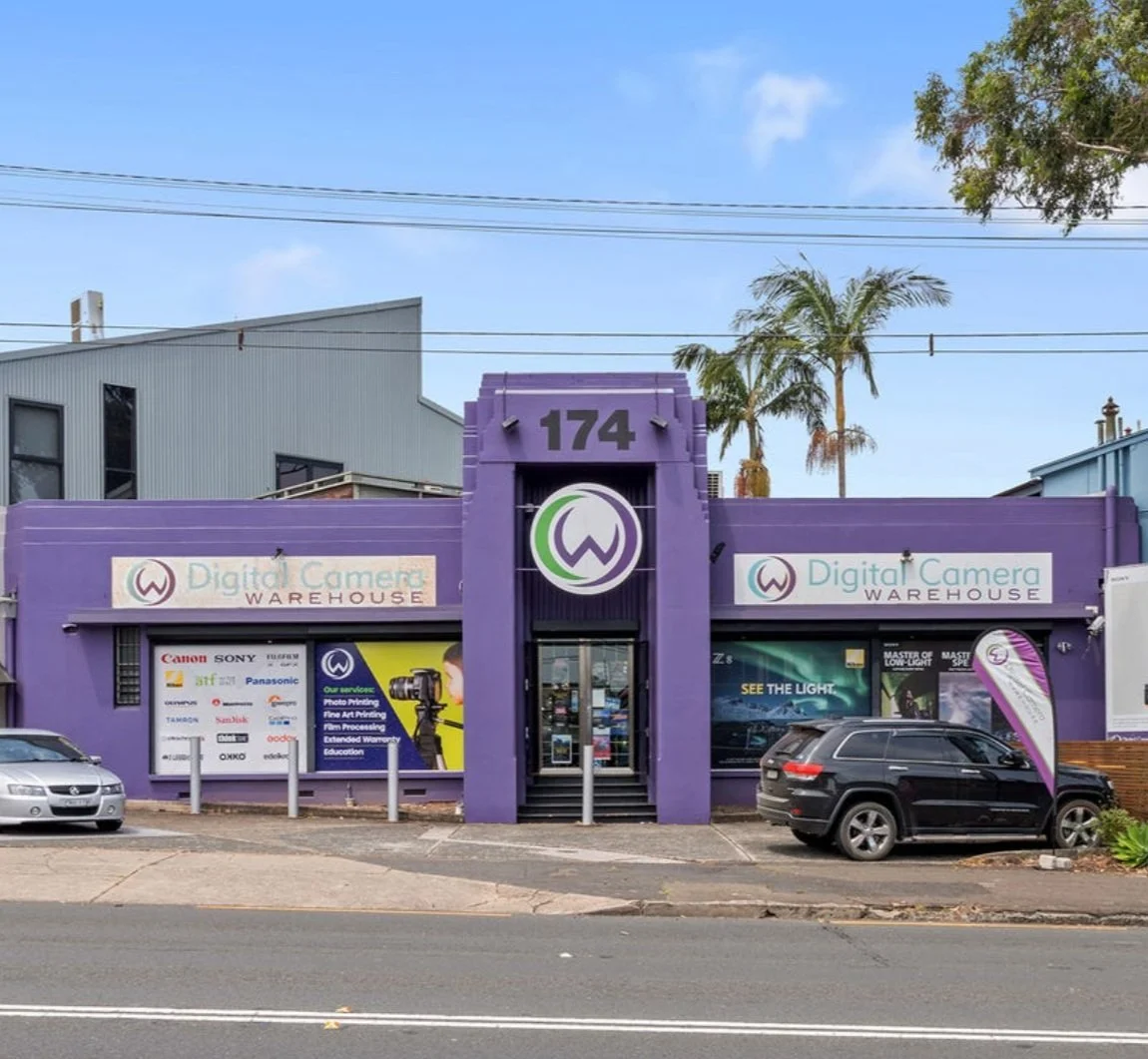 A purple building with a sign reading 'Digital Camera Warehouse' and a large purple logo above the entrance. Several cars are parked in front, and there are palm trees and a blue sky in the background.