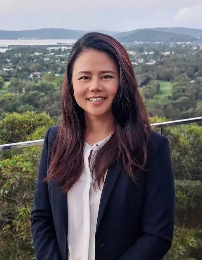 A woman with long dark hair, wearing a navy blazer and white blouse, smiling outdoors with a scenic landscape of trees, water, and hills in the background.