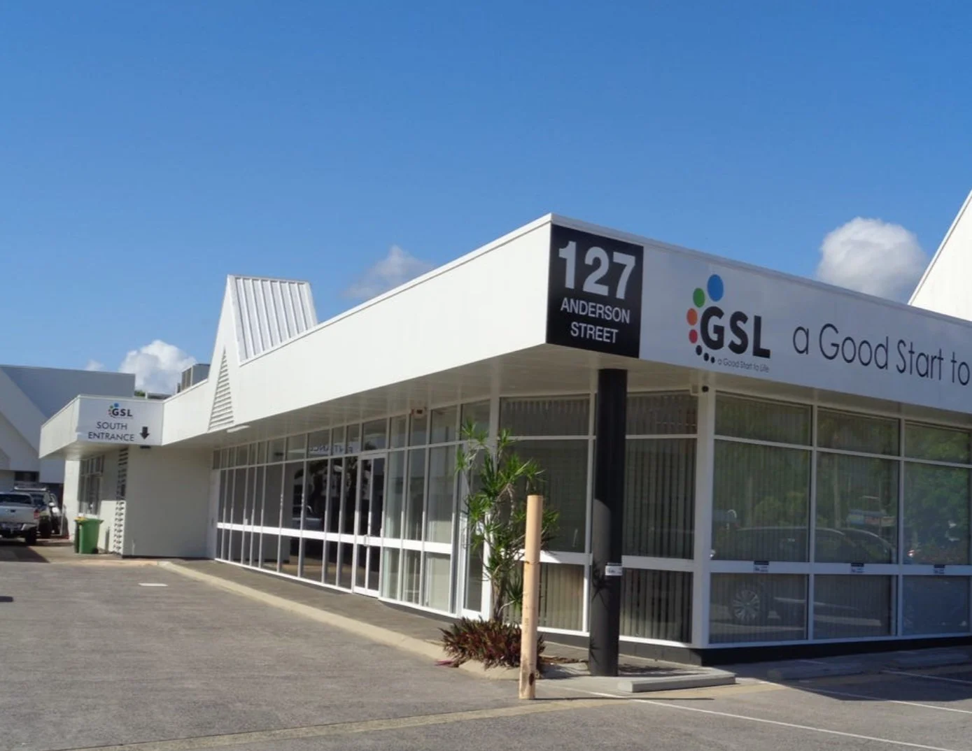 Exterior view of a GSL building with signage indicating its location at 127 Anderson Street, featuring modern white architecture with large glass windows, a parking lot, and a green trash bin under a partly cloudy sky.