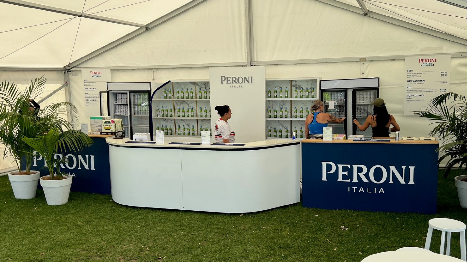 A Peroni Italia beverage stand inside a tent, with a white and navy blue counter displaying the Peroni logo, inside a grassy area with potted plants.