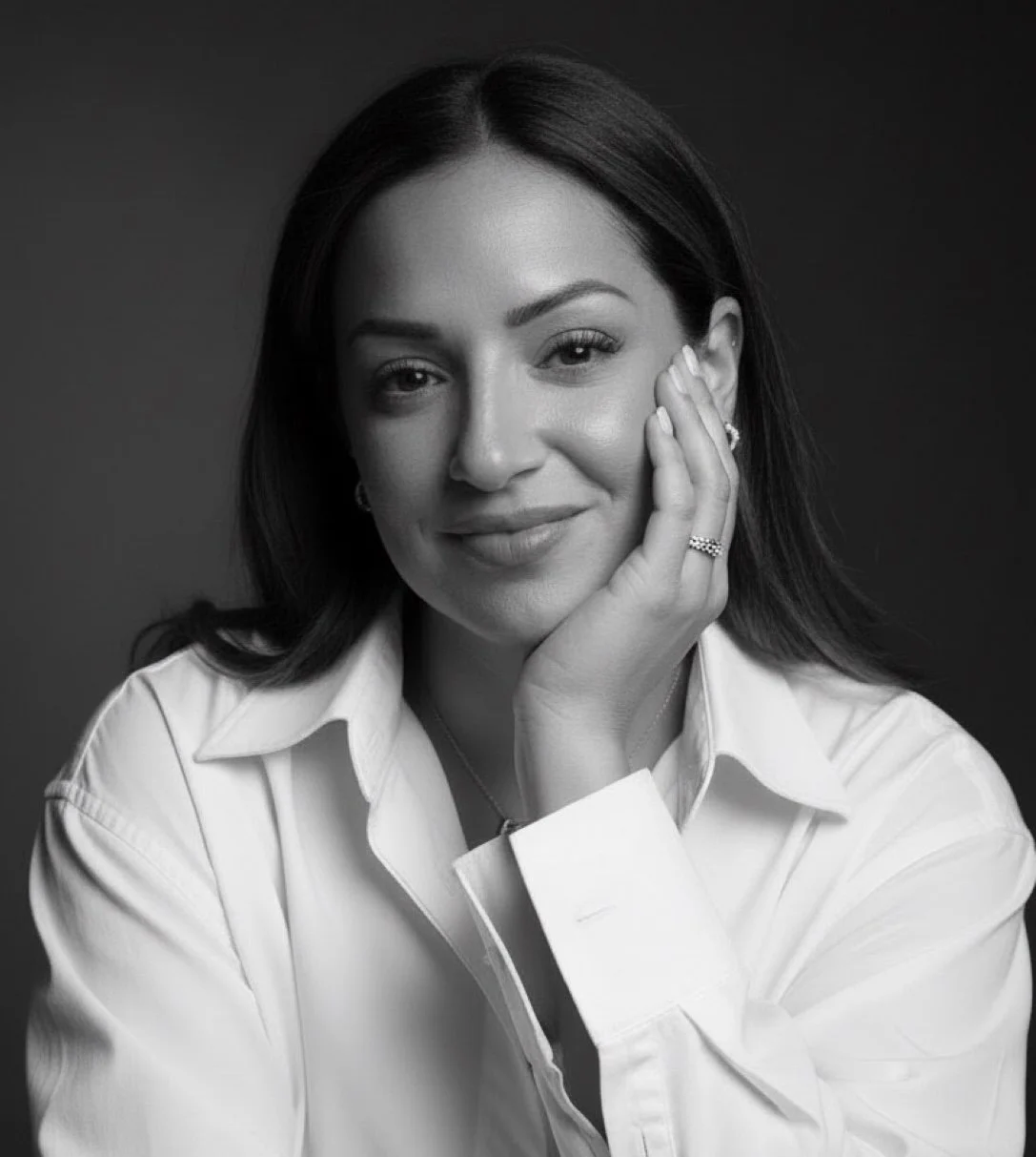 Black and white portrait of a woman with dark hair, resting her face on her hand, wearing a white shirt and jewelry, smiling softly.