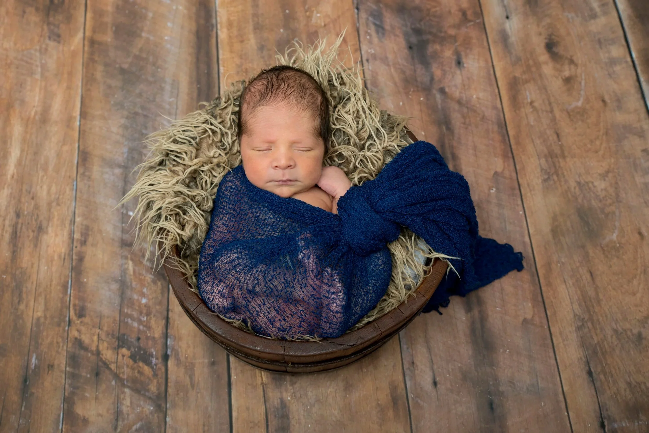 A newborn baby sleeping peacefully, wrapped in a dark blue cloth, and lying on a fluffy beige blanket inside a round wooden bed, with a rustic wooden floor visible underneath.