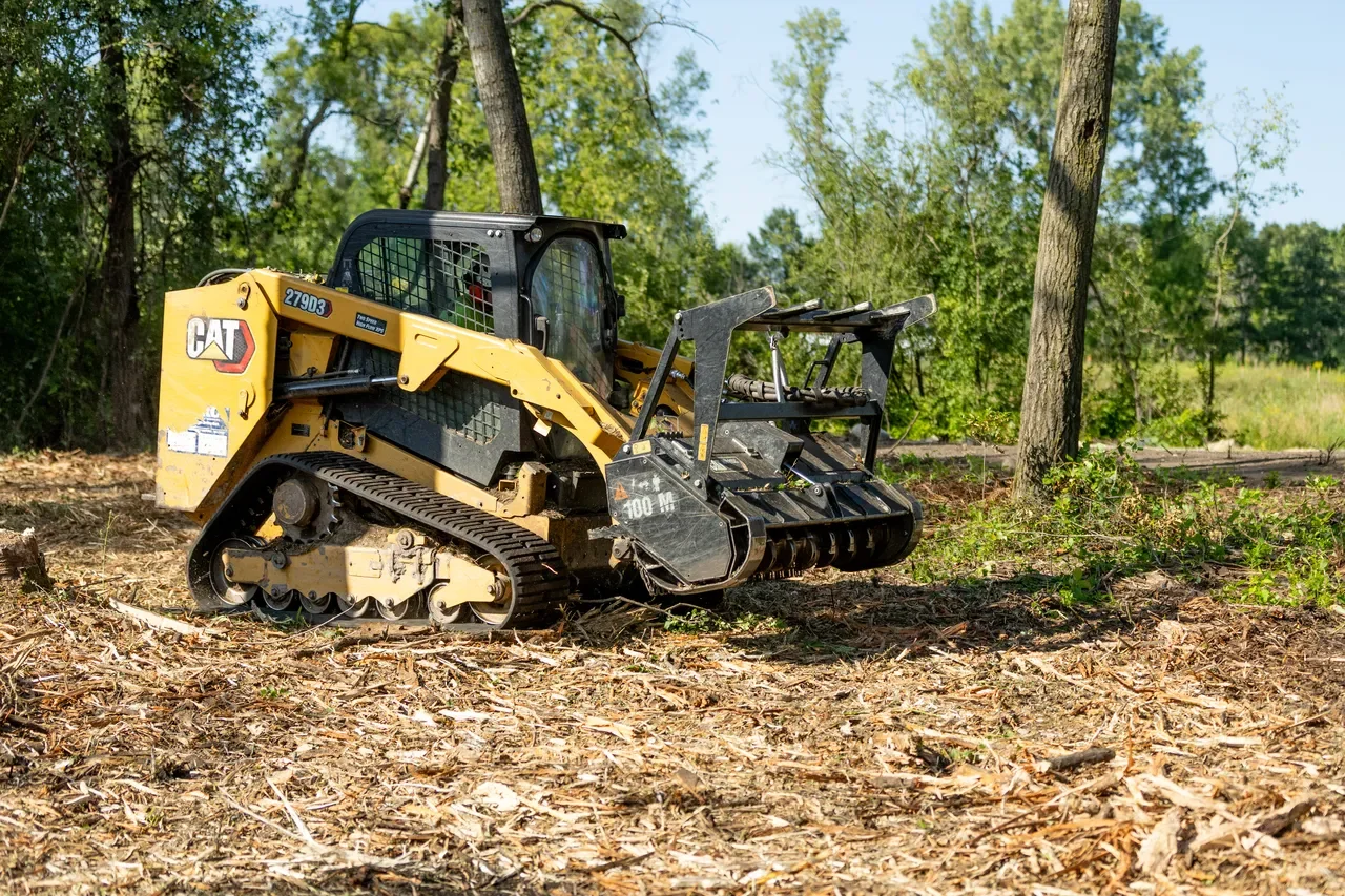 A bulldozer with caterpillar tracks in a wooded area. The background has trees and green foliage under a clear blue sky.