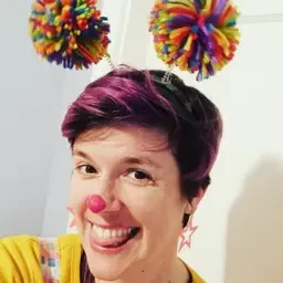 A woman smiling with pink hair, wearing star-shaped earrings, in front of colorful pom-poms hanging on the wall.