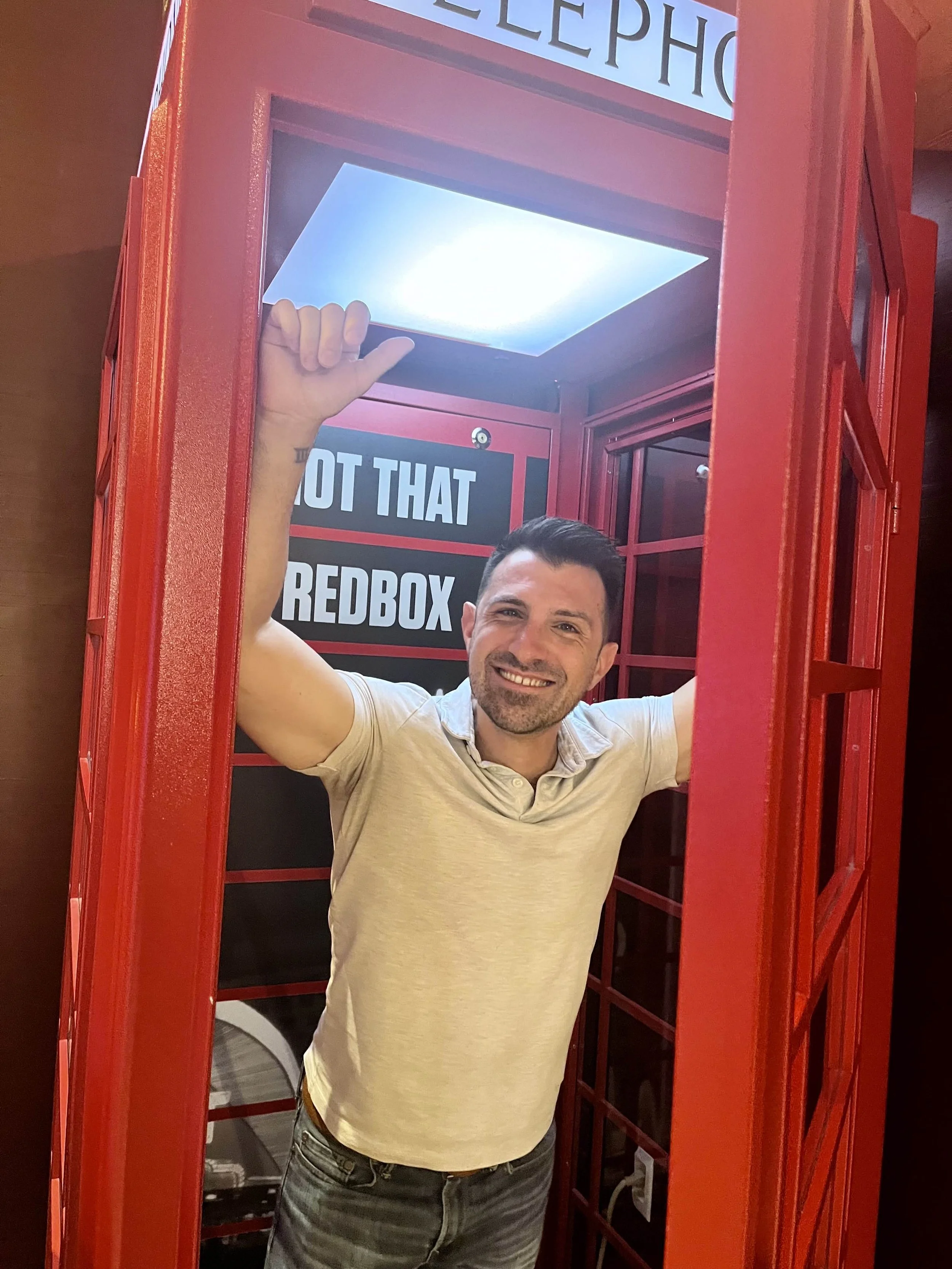 A man smiling inside a red British-style telephone booth, taking a selfie, with a sign that reads 'HOT THAT REDBOX' in the background.