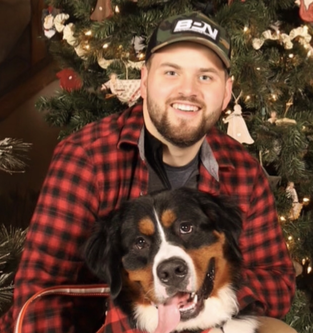 A smiling man wearing a camouflage baseball cap and a red plaid shirt sitting with a Bernese Mountain Dog in front of a decorated Christmas tree.