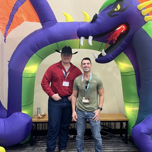 Two men standing inside a large purple and green inflatable dragon with yellow horns and red eyes at an indoor event.
