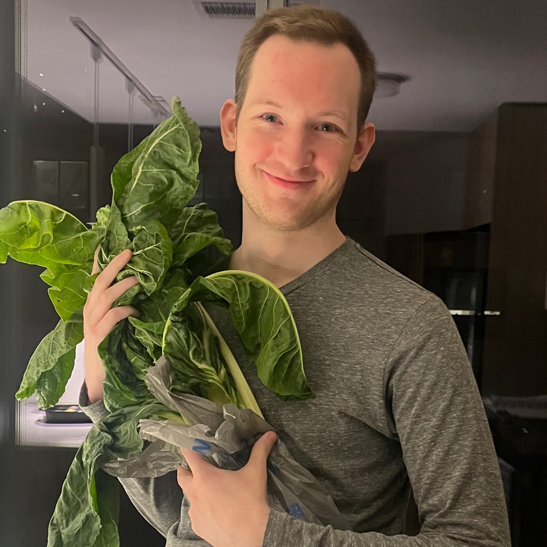 Young man with light brown hair holding a large leafy green plant while standing indoors.