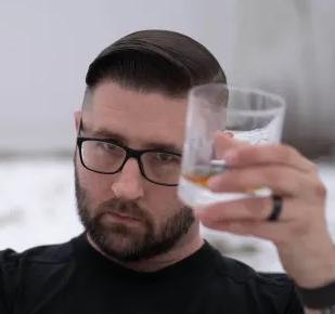 A man with dark hair and glasses raising a glass of whiskey or liquor.