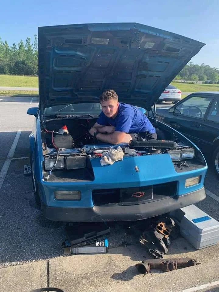 A man in a blue shirt working under the open hood of a blue vintage Chevrolet Corvette parked in a parking lot.